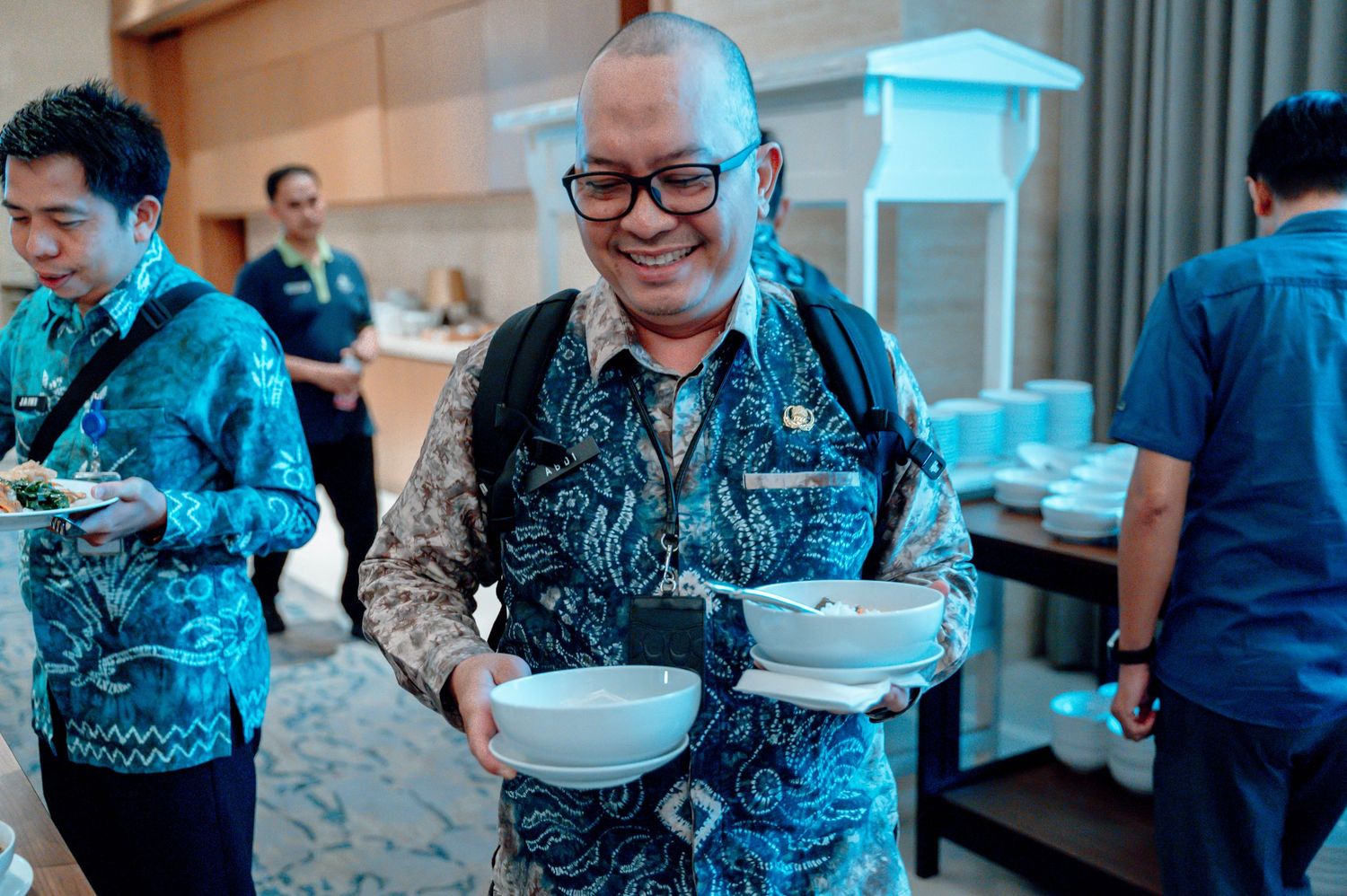 Man in batik shirt smiling while holding bowls at a buffet event.