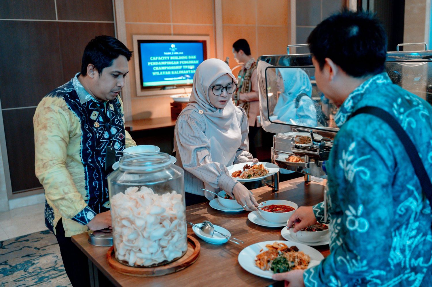 People in traditional batik attire serving food at an indoor buffet spread.
