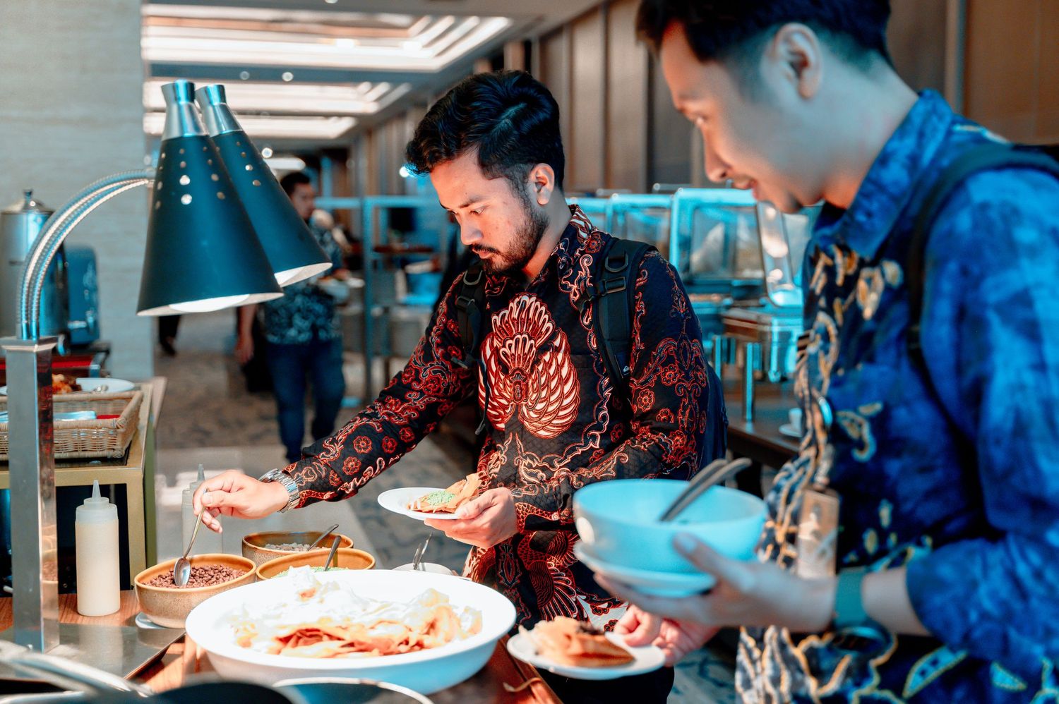 Two men in batik attire serving food at an elegant buffet spread in a upscale restaurant setting.