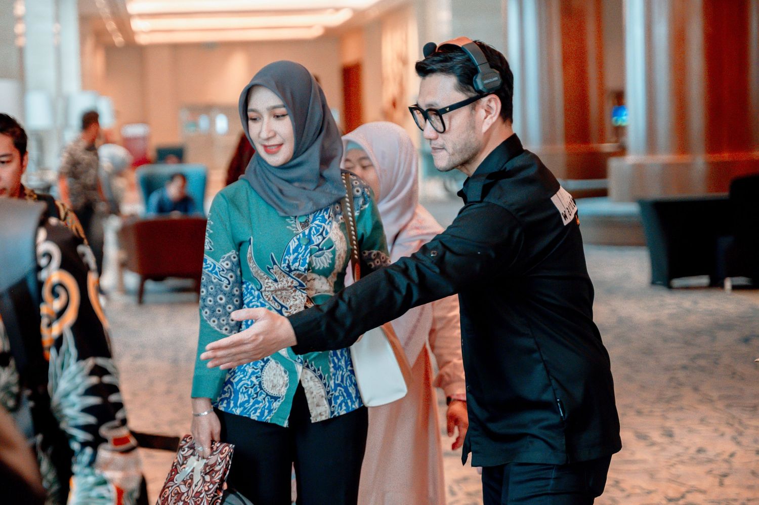 Man in black suit takes selfie with two women in hijabs and batik attire at an elegant indoor event.