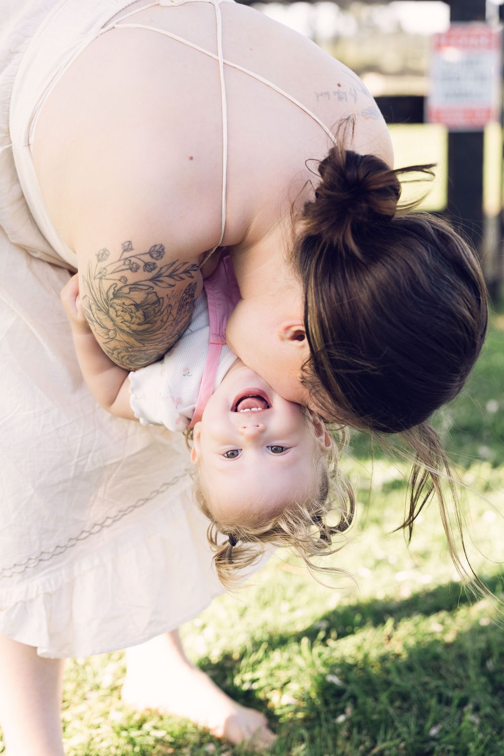 A tattooed mother playfully holds her laughing toddler upside down on a sunny green lawn.