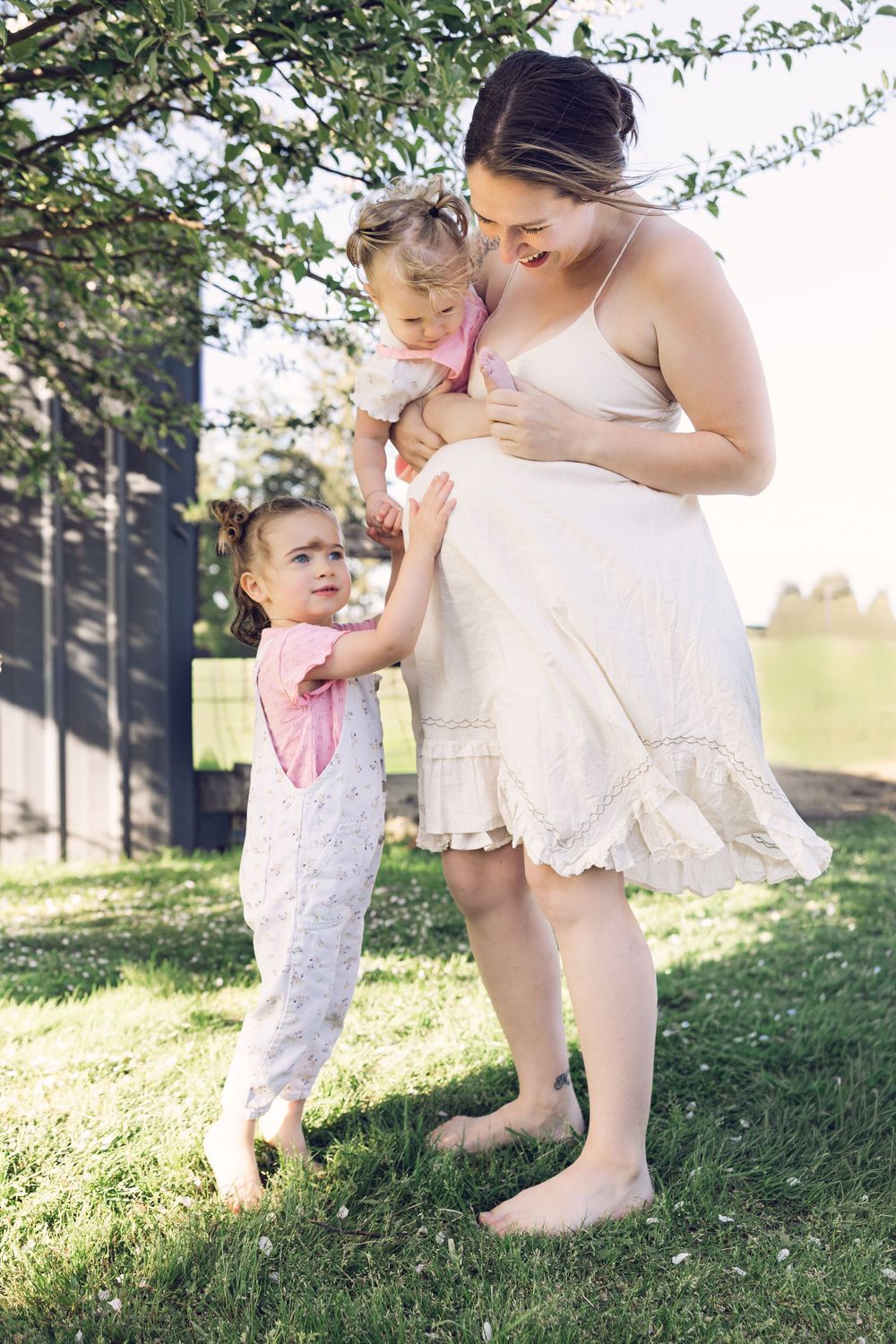 Pregnant mother holding toddler while young daughter stands beside her outdoors on green grass.
