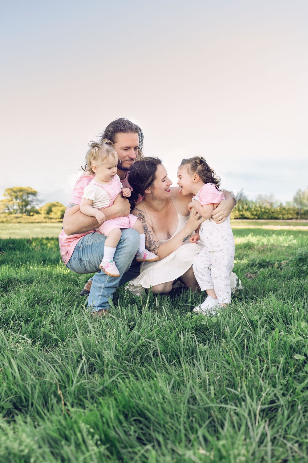 Happy family with two toddler girls sitting on green grass in a sunny park, parents smiling and embracing children.