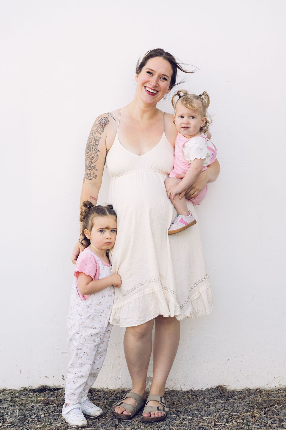 Pregnant tattooed mother in white dress smiling while holding two young daughters against white wall.
