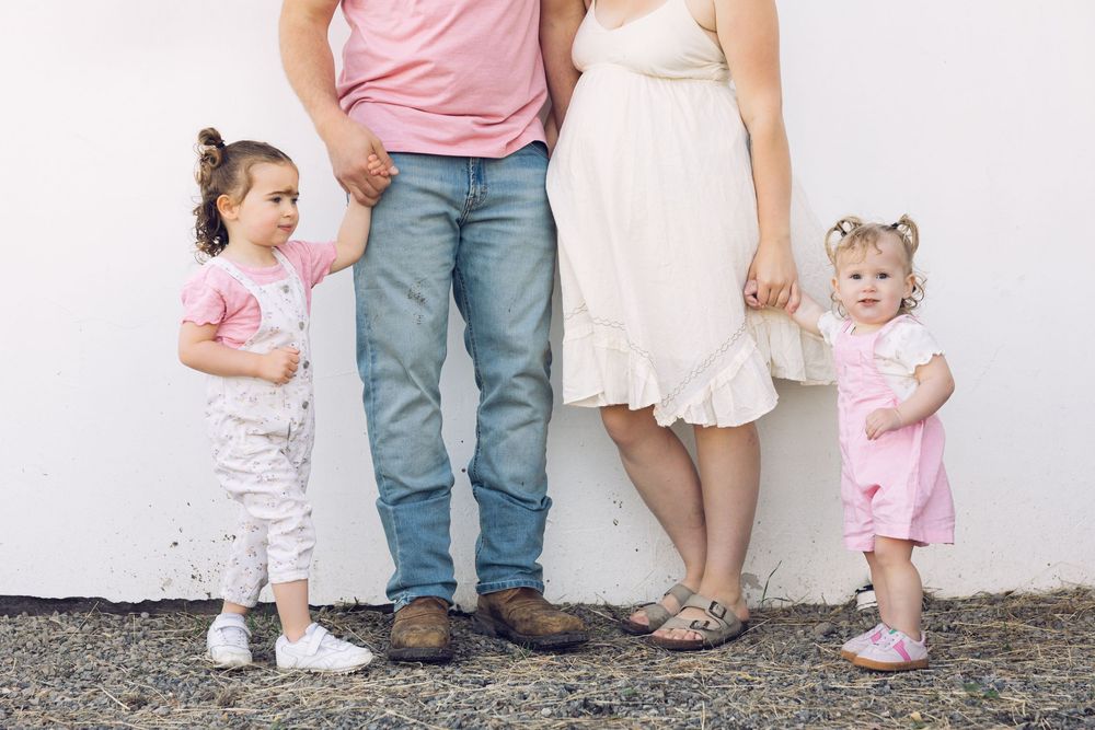 A family of four with two young girls in pink outfits stand against a white wall holding hands.