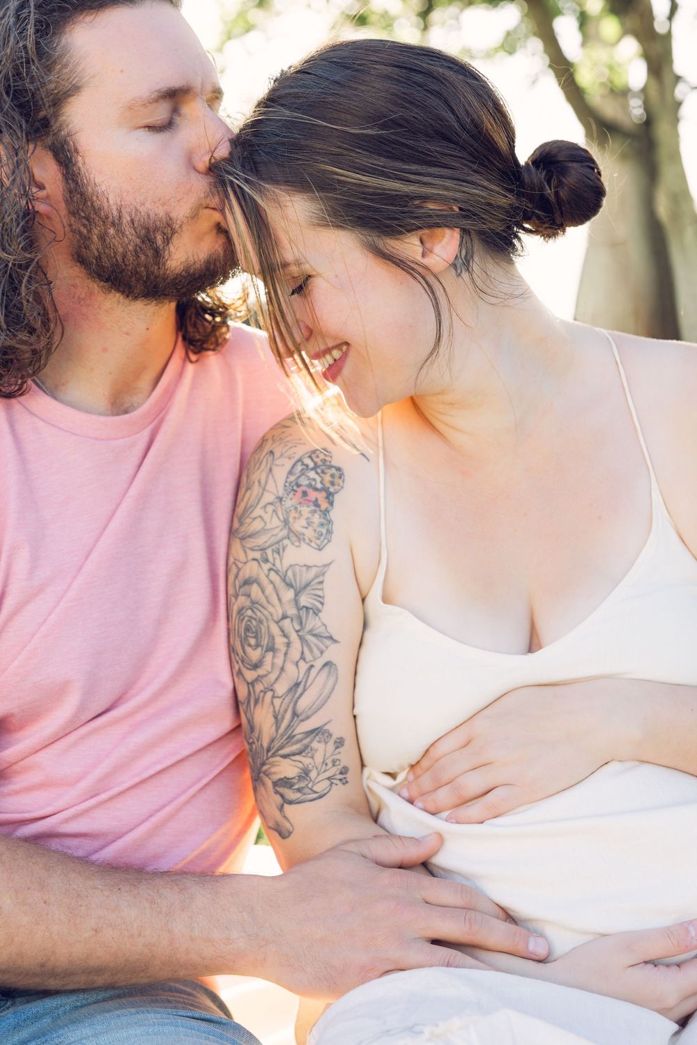 Bearded man in pink shirt kissing smiling pregnant tattooed woman outdoors in warm sunlight.