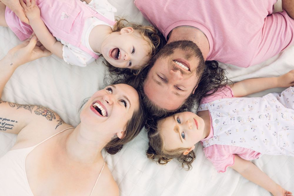Happy family of four lying in bed together laughing, wearing pink pajamas in a cozy overhead portrait.