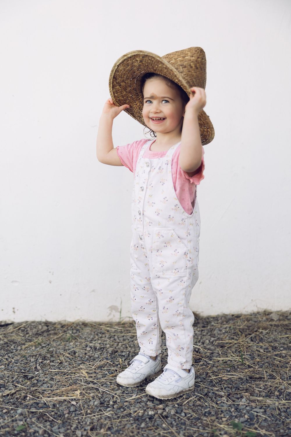 Adorable toddler girl wearing oversized straw hat in white floral overalls smiling against white wall.