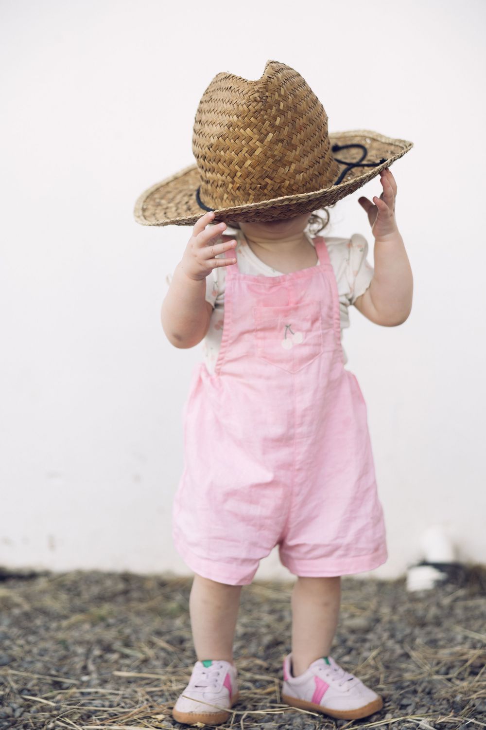 Toddler in pink overalls and sneakers wearing an oversized straw cowboy hat covering her face.