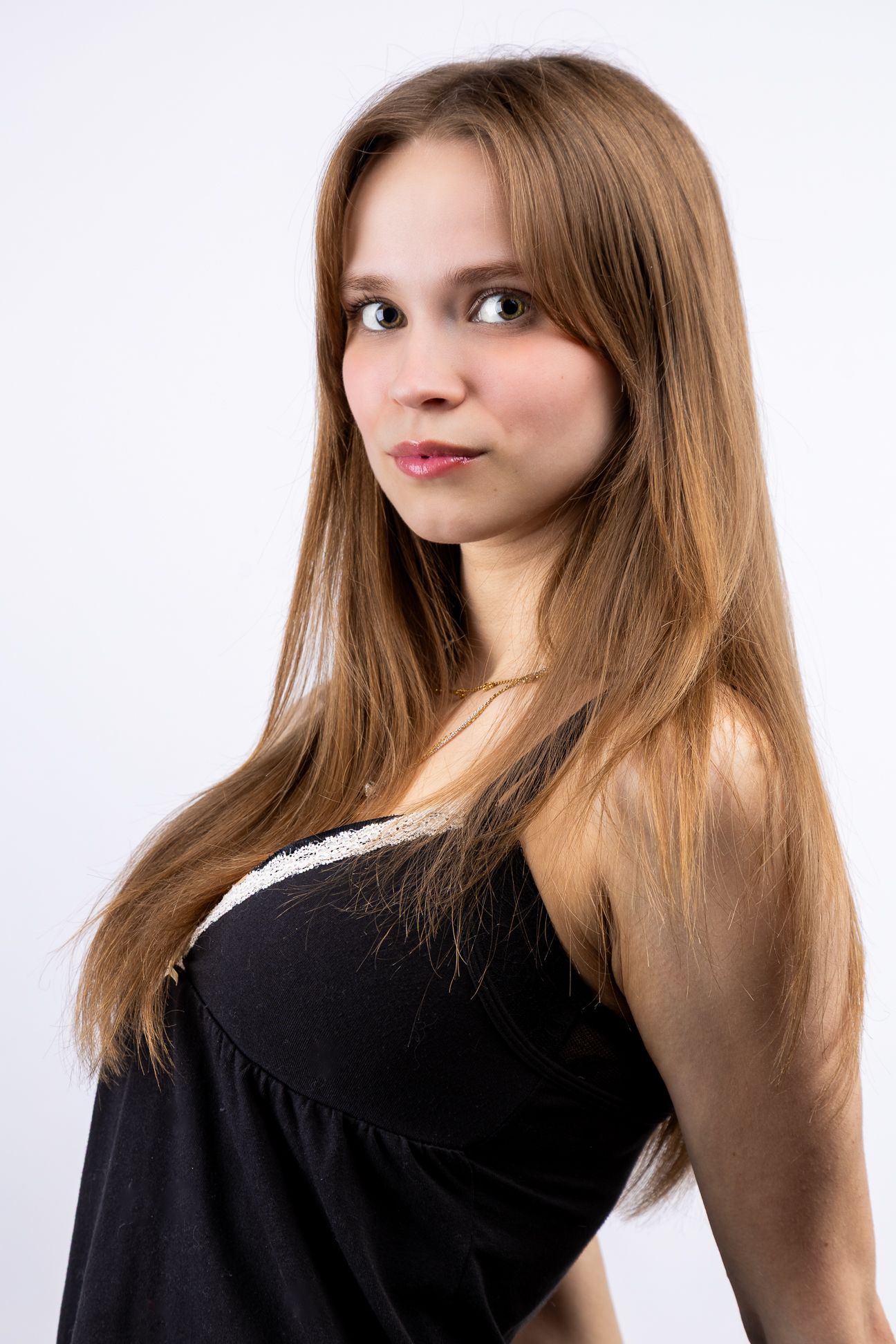 A girl in front of a white backdrop in a black tank top, looking at the camera with arms back