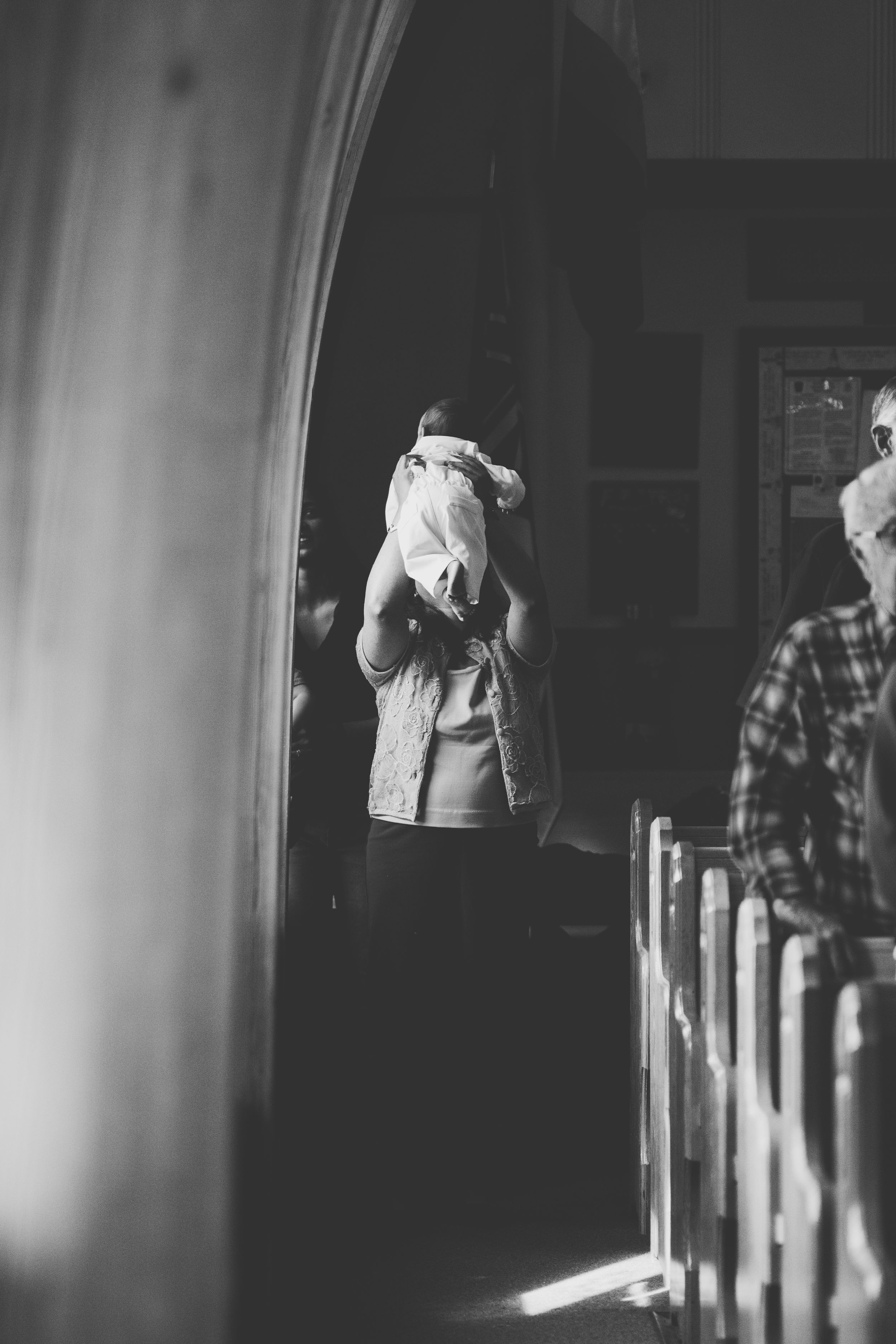 Black and white photo of a person holding a baby inside a church with pews and an arched doorway.