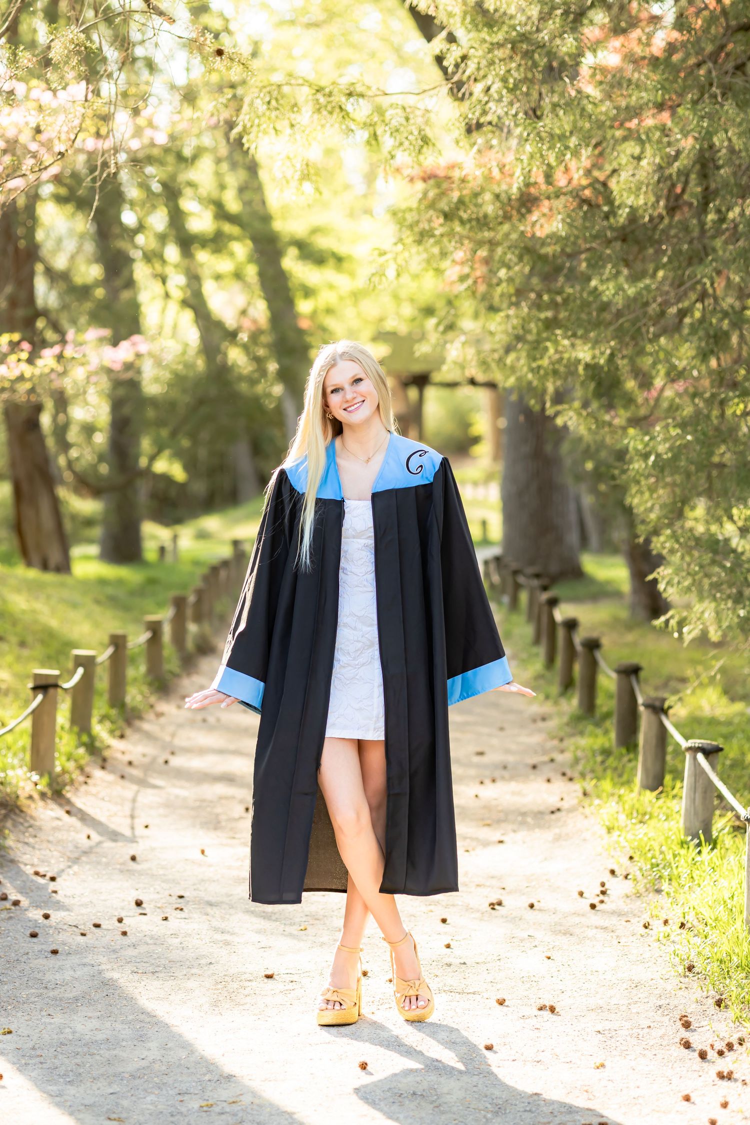 Smiling blonde woman in black and blue graduation gown posing on a sunlit tree-lined path.