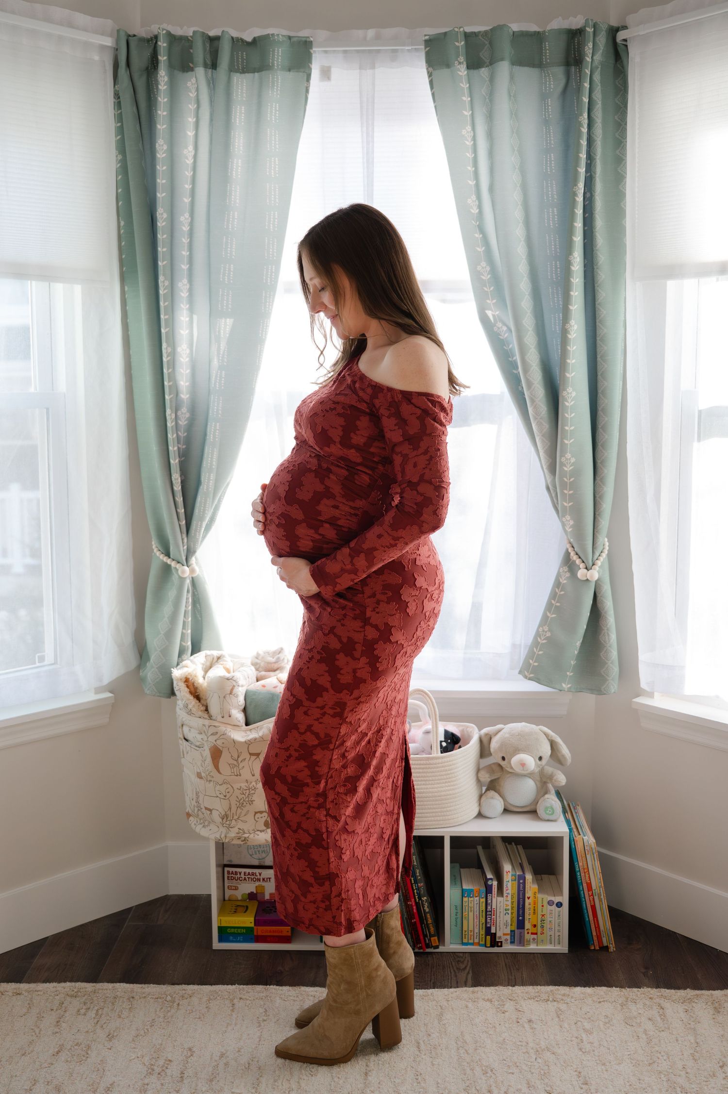 Pregnant woman in red floral dress and tan boots standing by window in nursery, cradling baby bump during maternity session, Rockland County