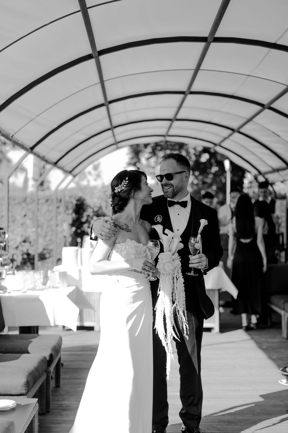Black and white photo of a smiling bride and groom toasting with champagne under a curved canopy at an outdoor wedding reception.