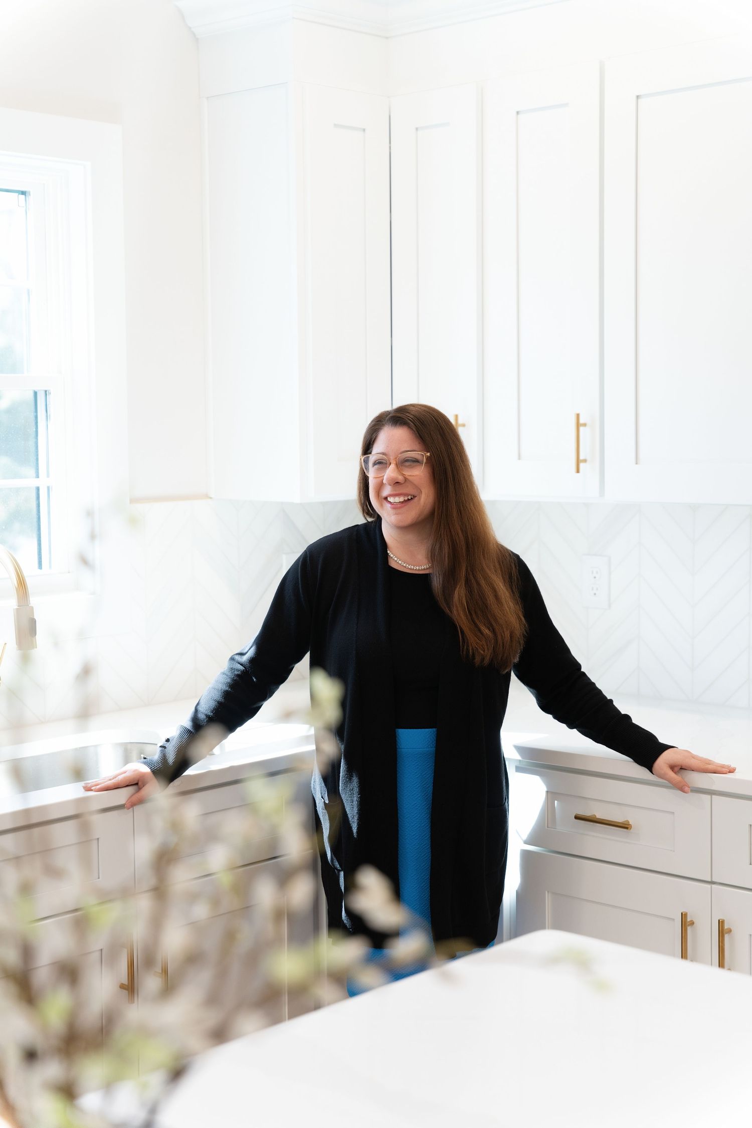 Woman smiling in a bright white kitchen with shaker cabinets, gold hardware, and a herringbone tile backsplash.