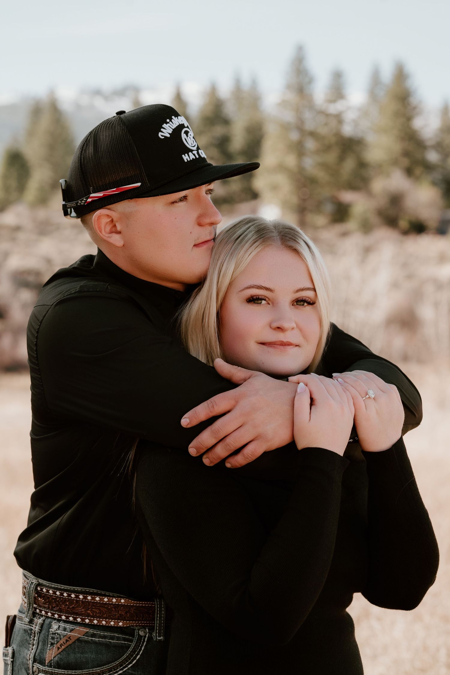 Couple in matching black outfits embracing in a field, man hugging woman from behind near pine trees and mountains.