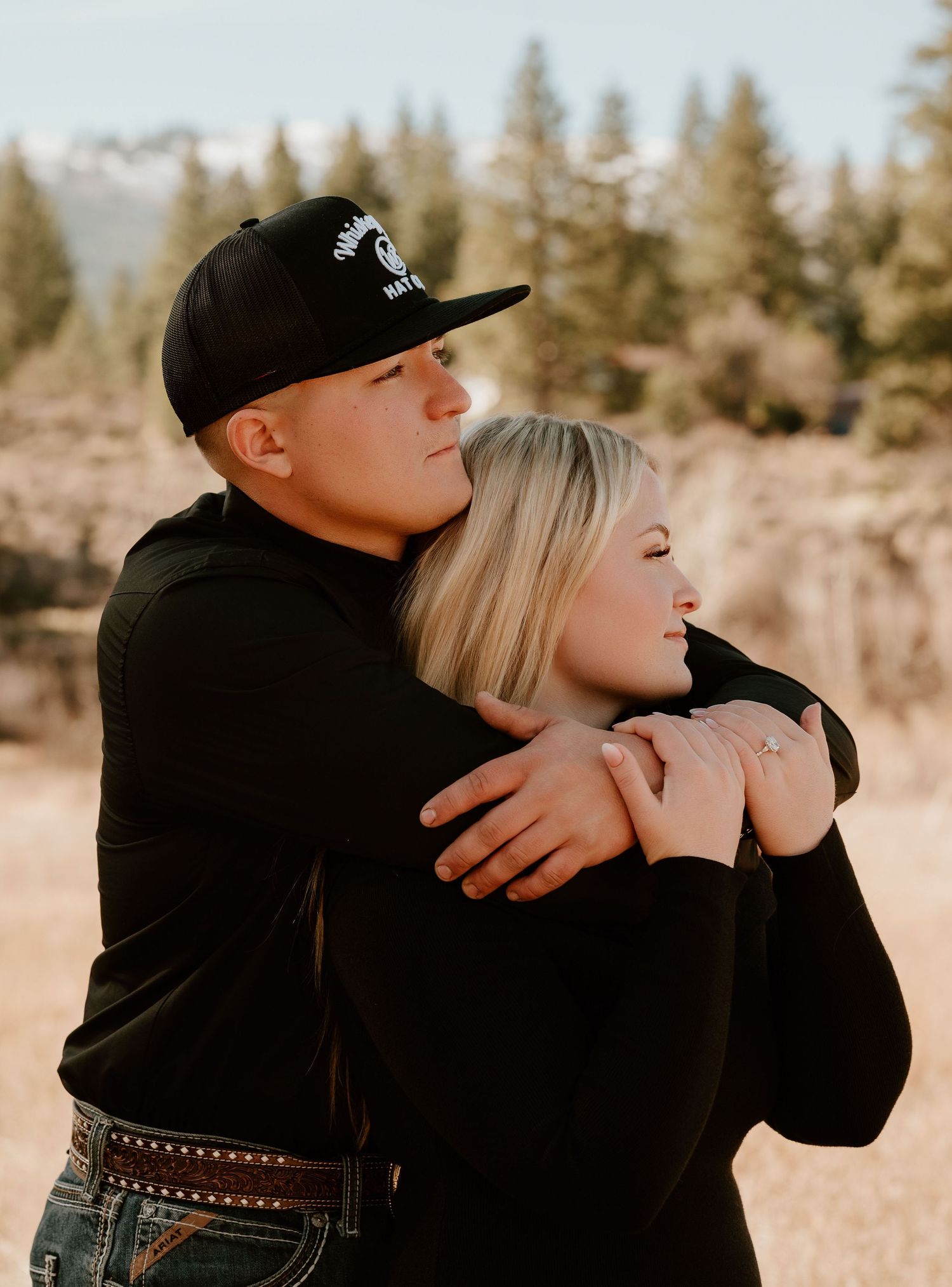 A couple embraces outdoors near pine trees, wearing matching black outfits, with mountains visible in the background.
