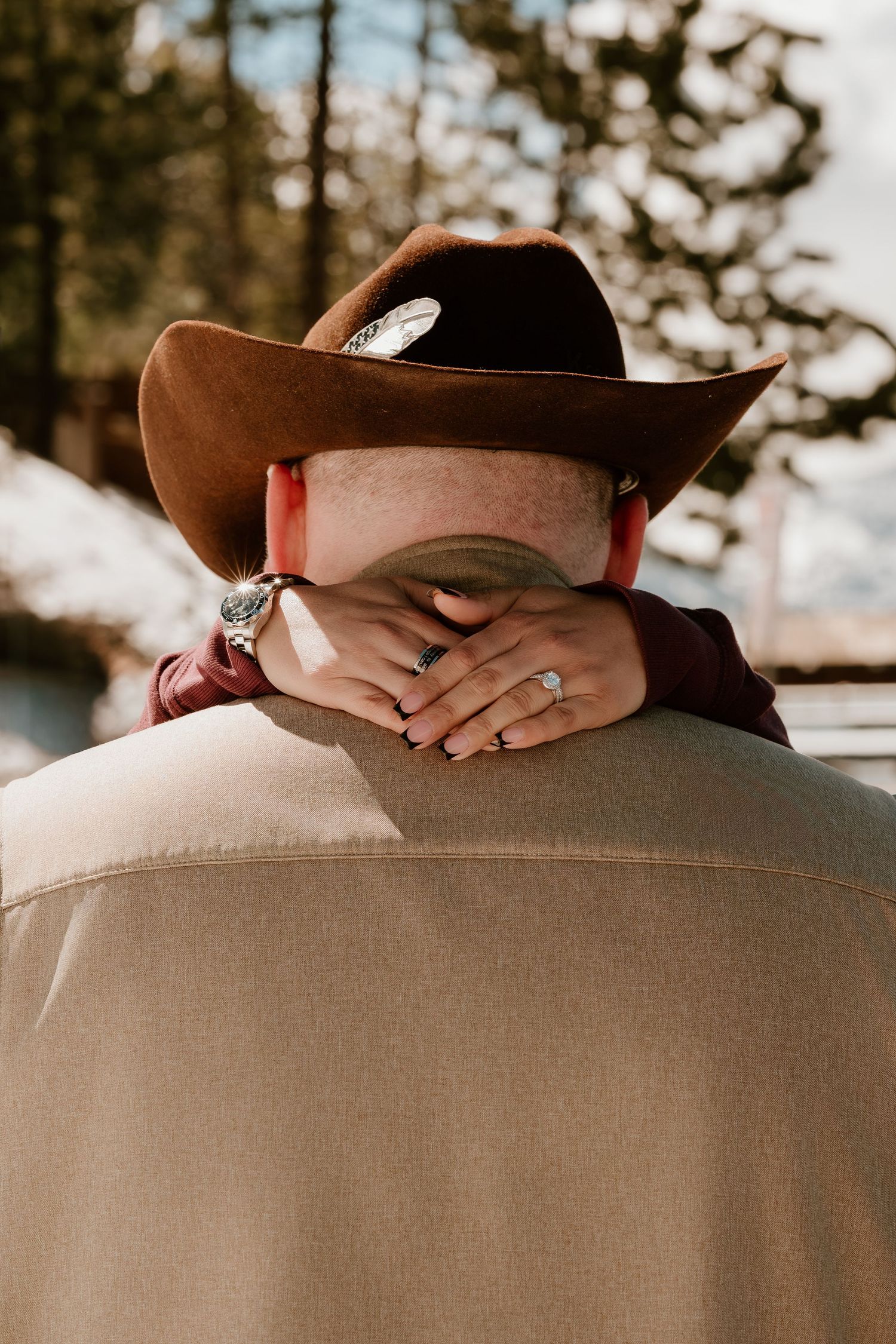 Couple embracing outdoors, man wearing brown cowboy hat with feather, woman's hands showing engagement ring.