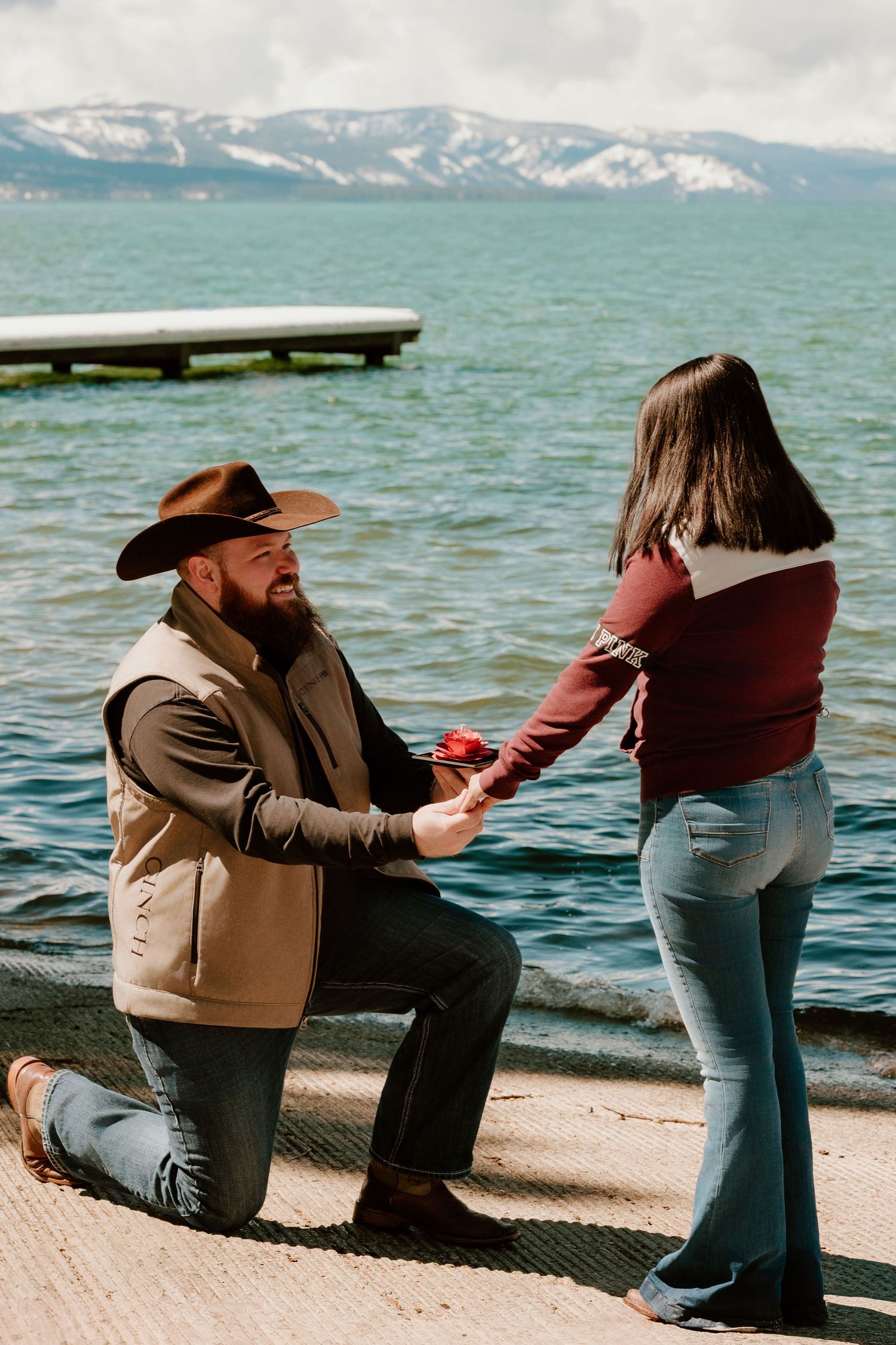 Man in cowboy hat proposes on one knee by a lake with snowy mountains, holding a red rose for his partner.