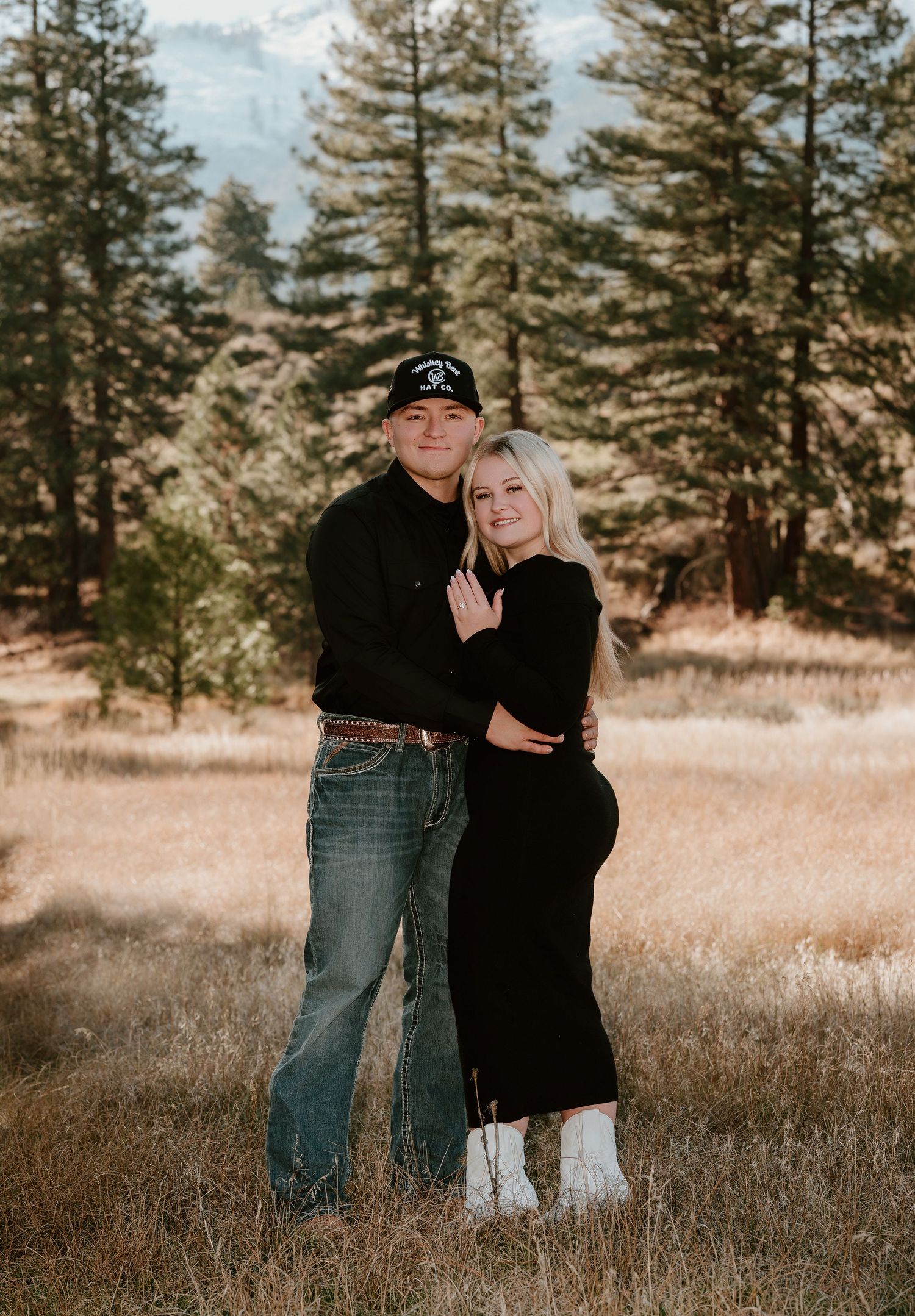 Engaged couple posing in a golden field wearing black outfits, woman showing ring near tall pine trees.