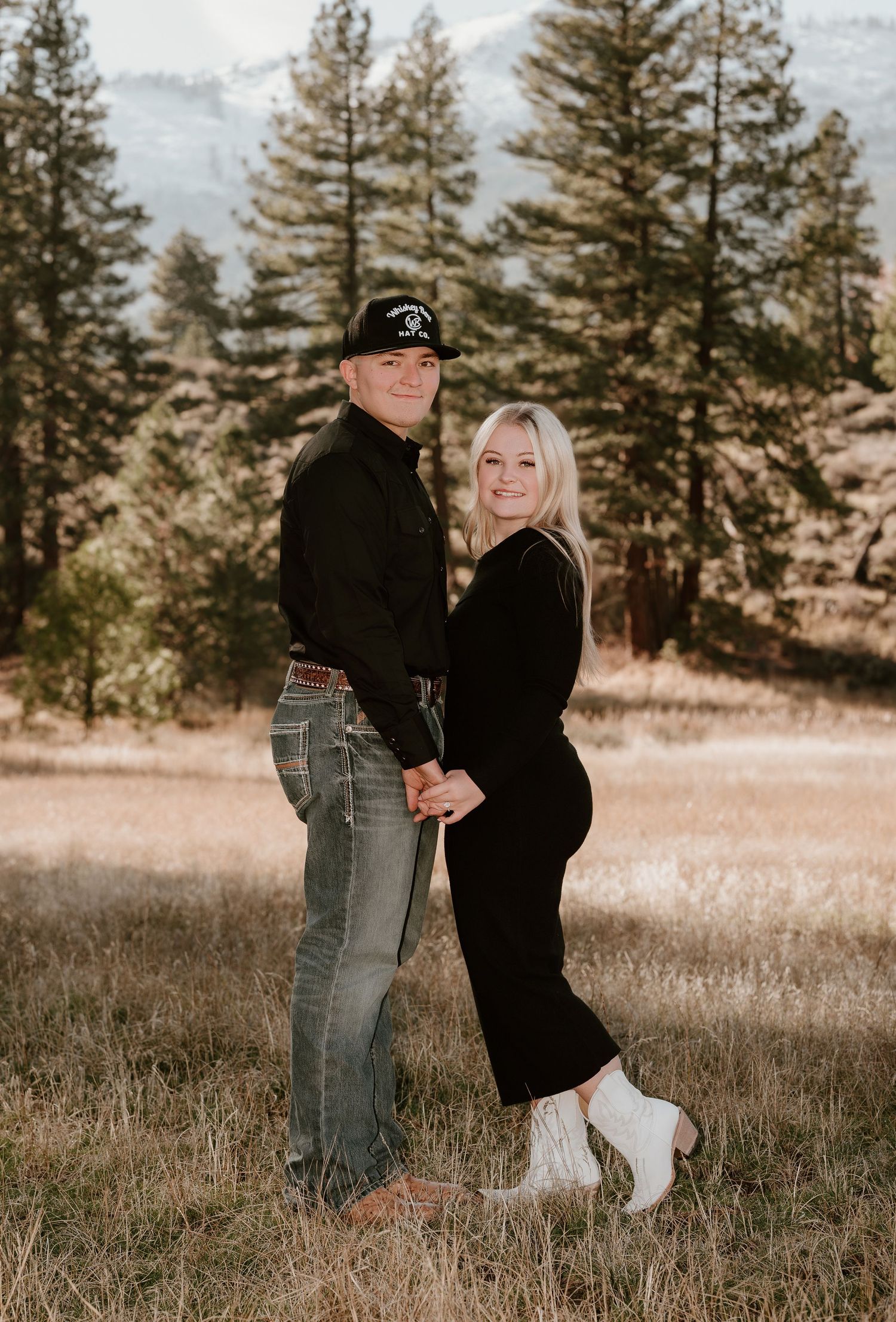 Couple holding hands in a mountain meadow, woman in black dress and white boots, man in jeans and black cap.