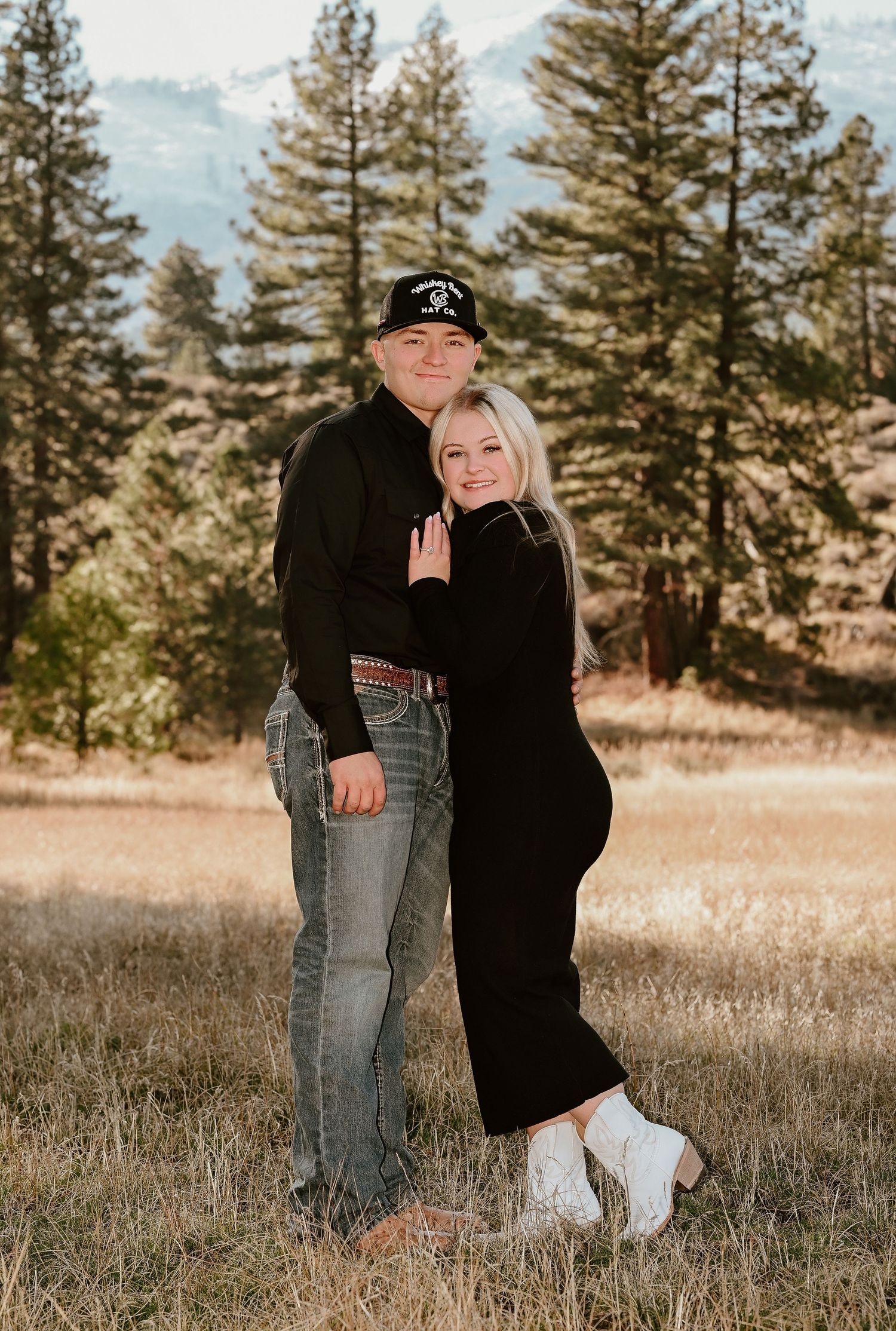 Couple embracing in a rustic mountain field, both dressed in black, surrounded by tall pine trees.