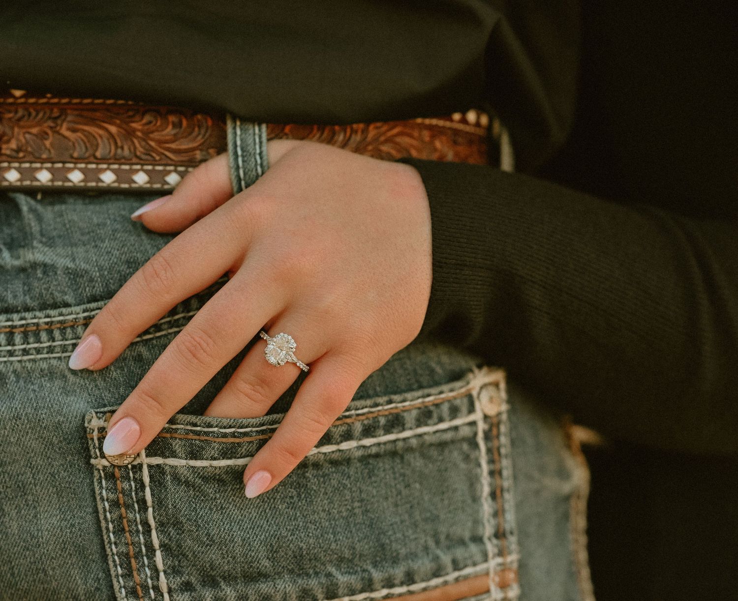 Close-up of a diamond engagement ring on a woman's hand tucked into a denim jeans pocket with a leather belt.