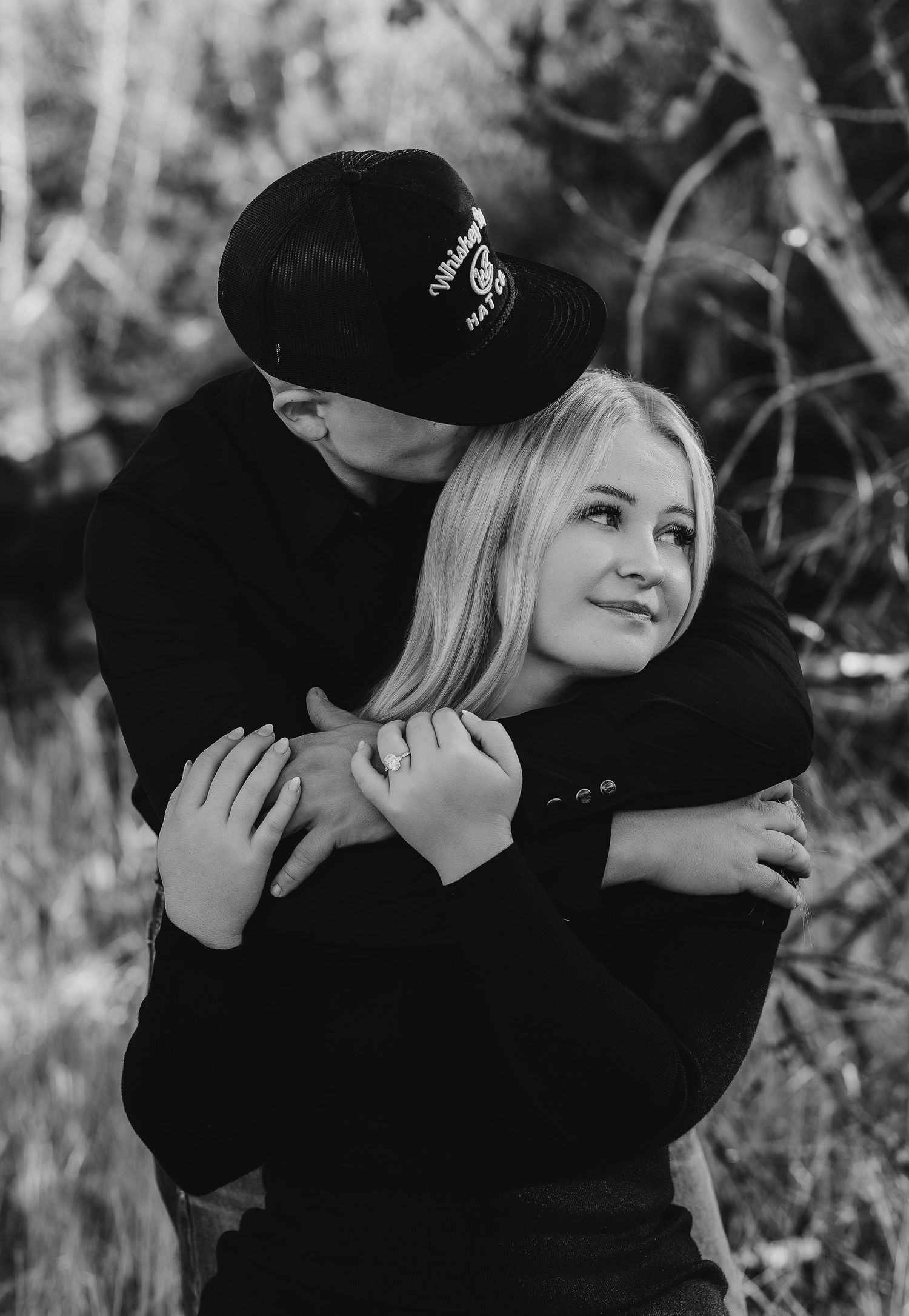 Black and white photo of a couple embracing outdoors, man in cap hugging woman from behind as she smiles warmly.