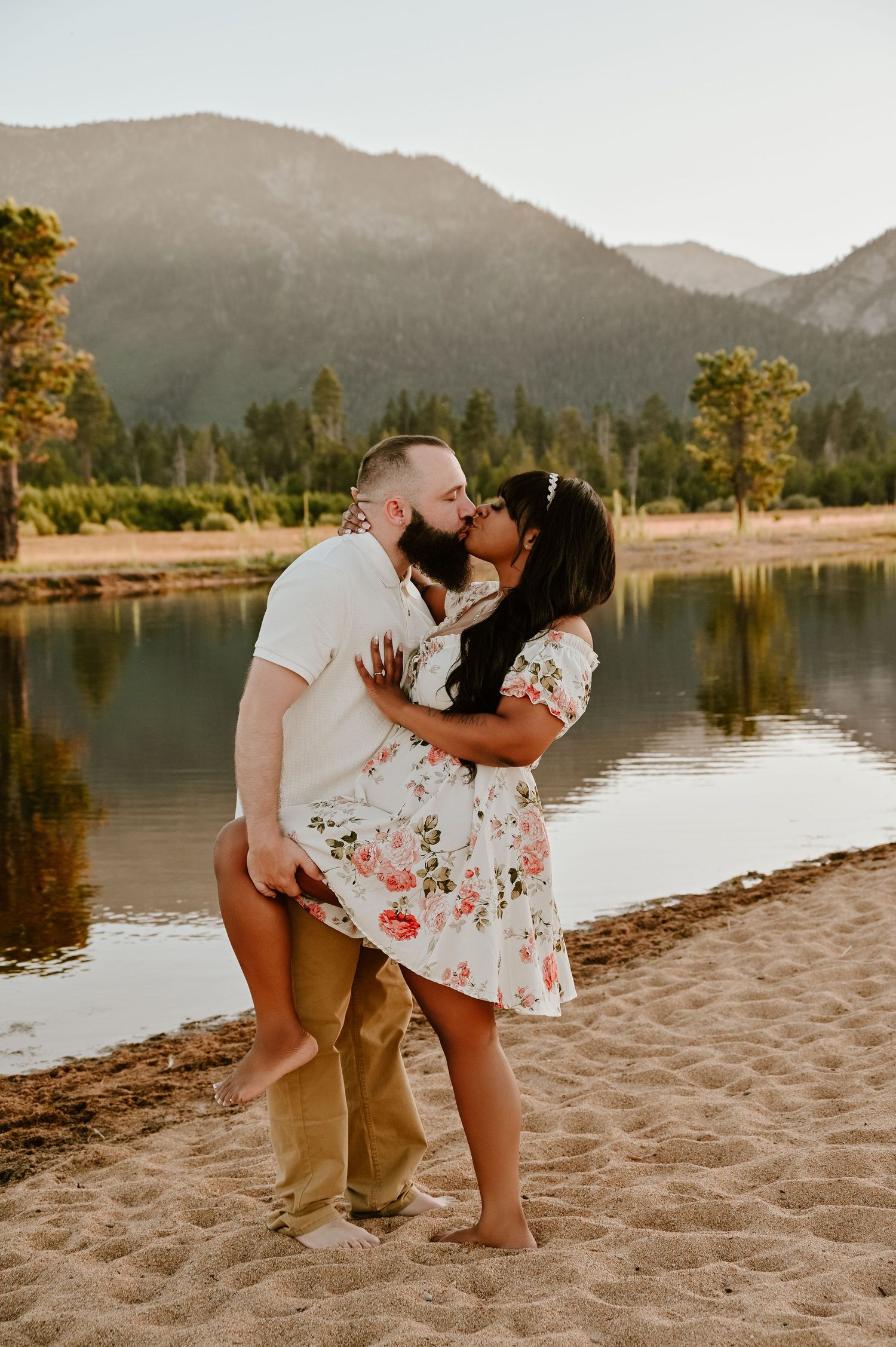 Couple sharing a romantic kiss on a sandy beach with a mountain lake and pine trees in the background.