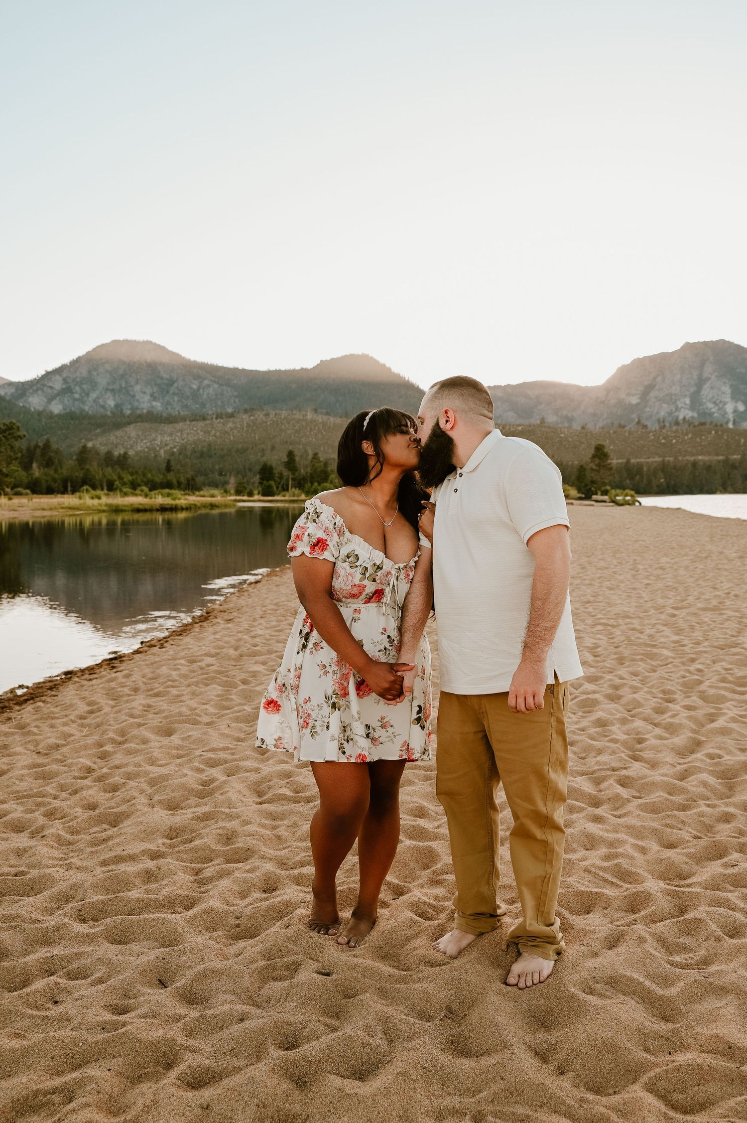 Couple sharing a kiss on a sandy beach with a serene mountain lake and trees in the background.