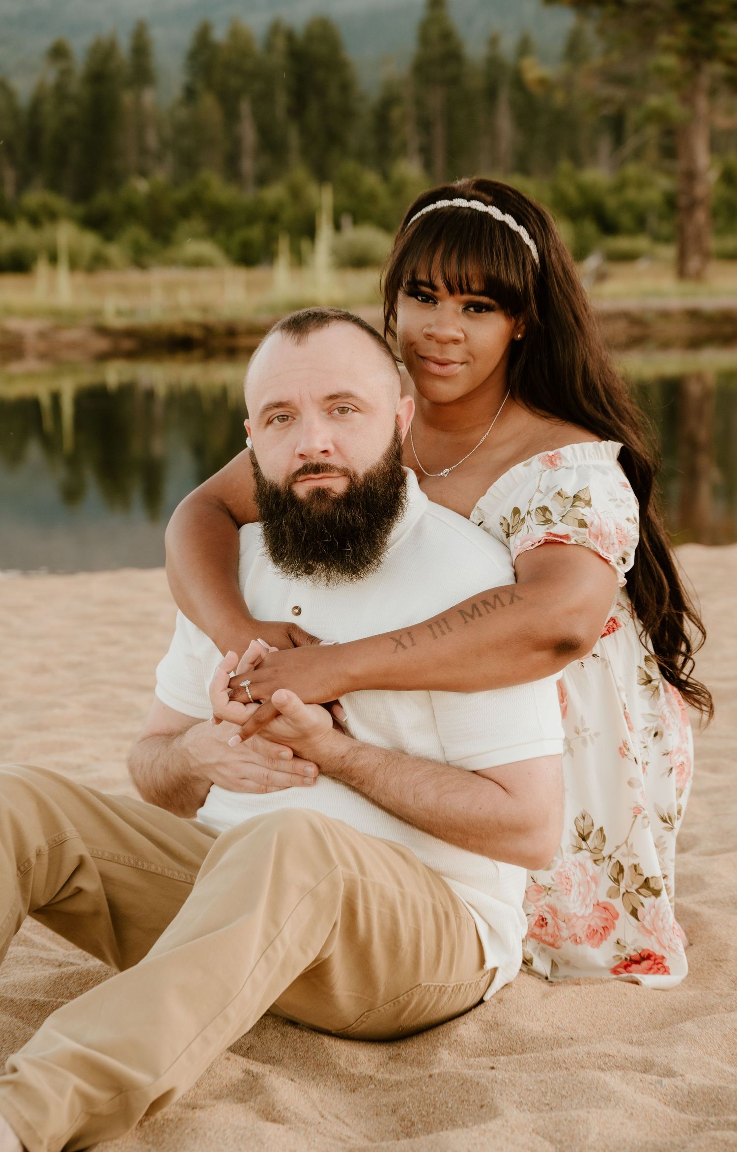 Couple posing on sandy beach by a lake, woman in floral dress embracing bearded man in white shirt.