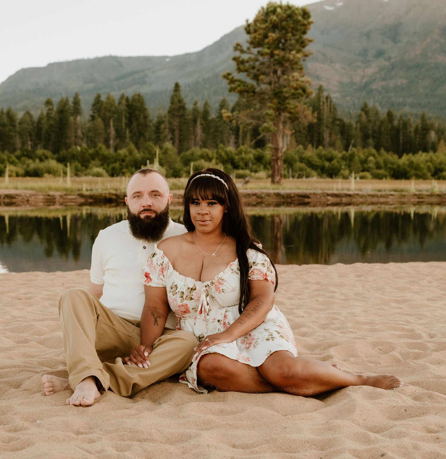 Couple sitting together on sandy beach with mountain lake and pine trees backdrop in scenic outdoor photo.