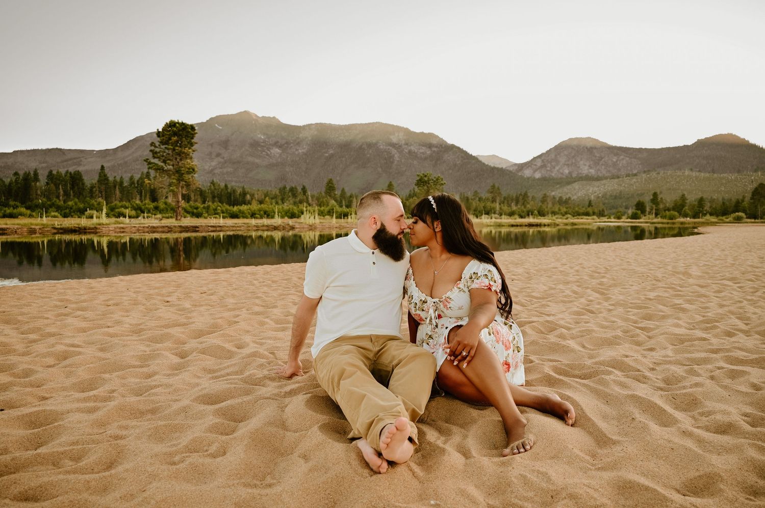 Couple kissing on sandy beach with mountain lake and pine trees backdrop during golden hour outdoor engagement session.