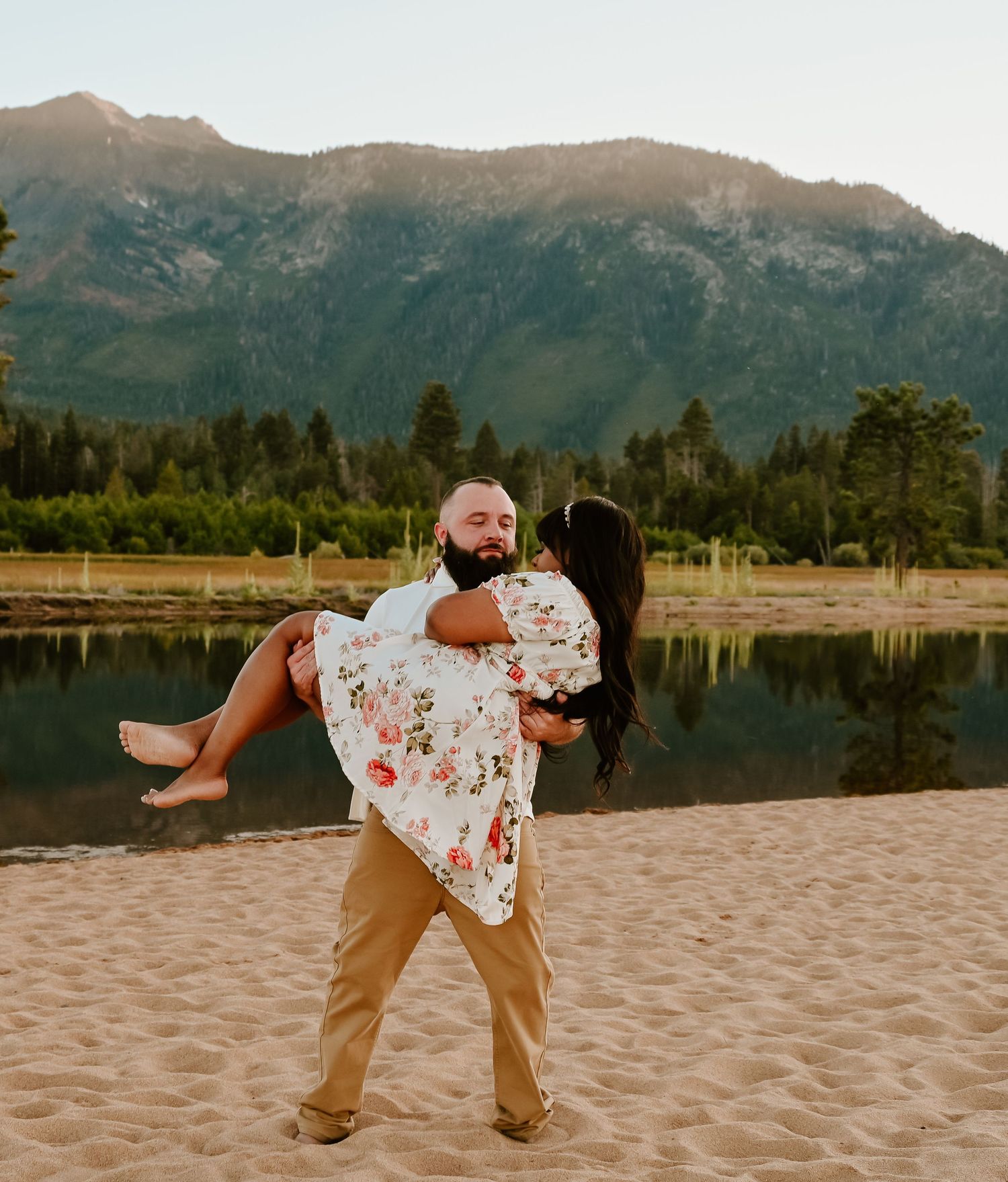 A bearded man carries a woman in a floral dress on a sandy beach with a mountain lake and forest backdrop.