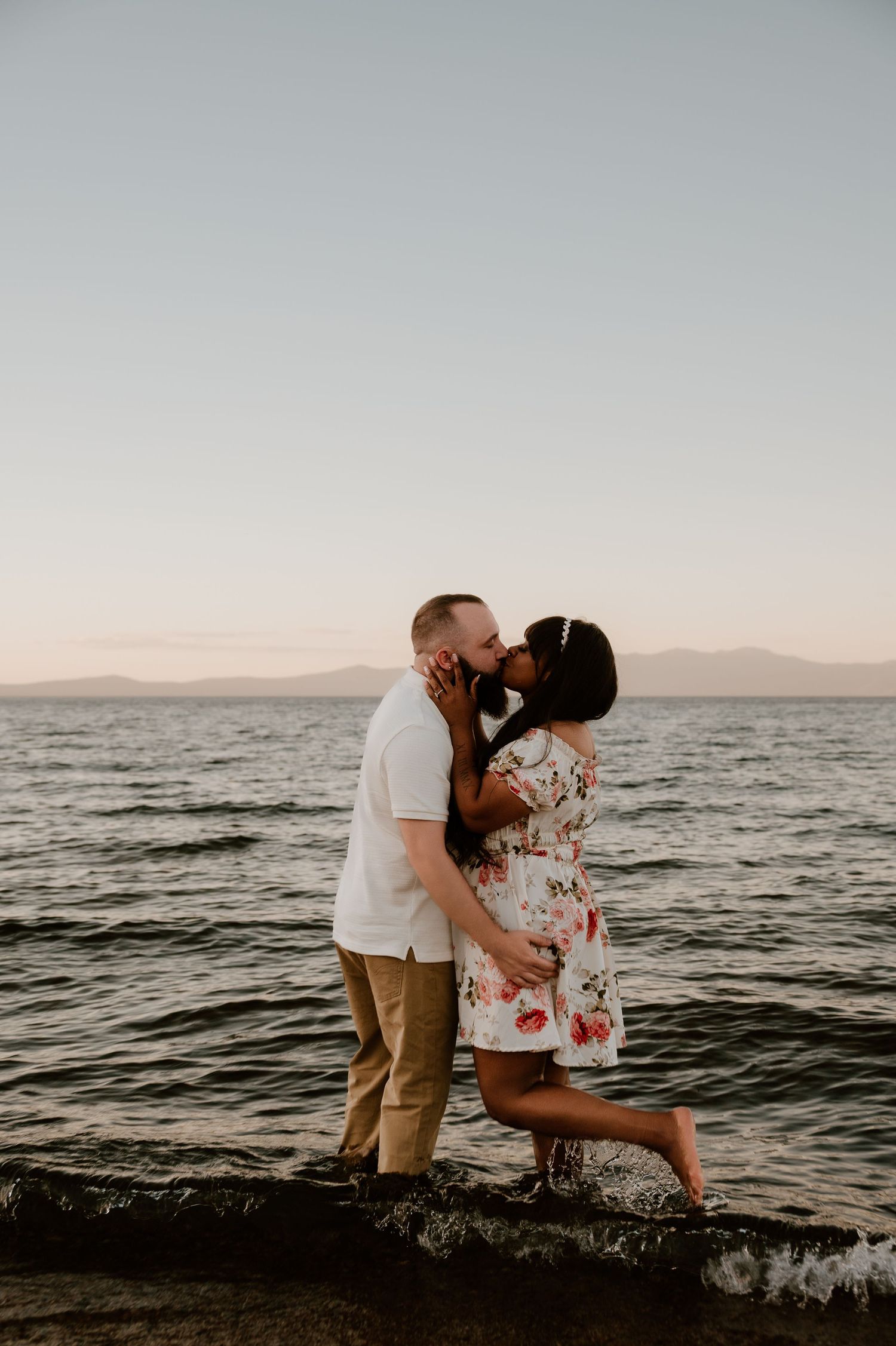 Couple sharing a romantic kiss by the lake at sunset, woman in floral dress standing on one leg near the water.
