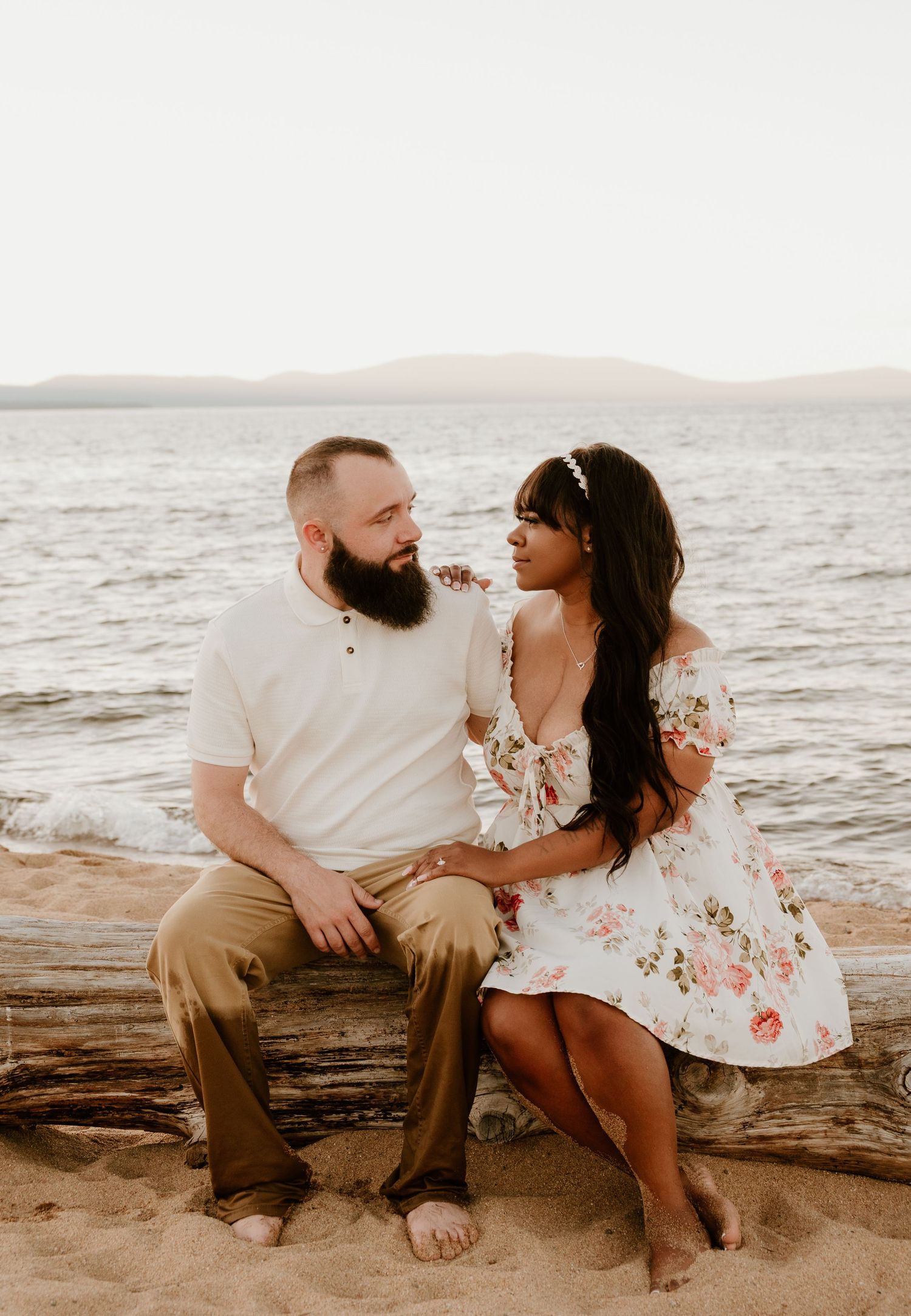 Couple sitting on a driftwood log on a sandy beach, gazing lovingly at each other with a calm lake behind them.