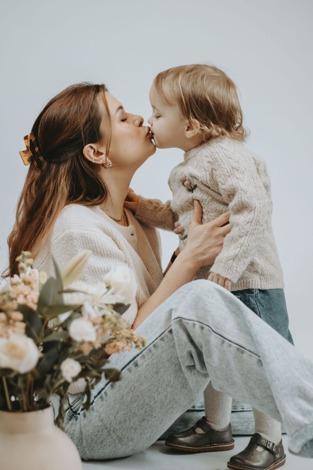 A young mother and toddler share a sweet kiss in a bright studio, surrounded by soft florals and cozy knitwear.