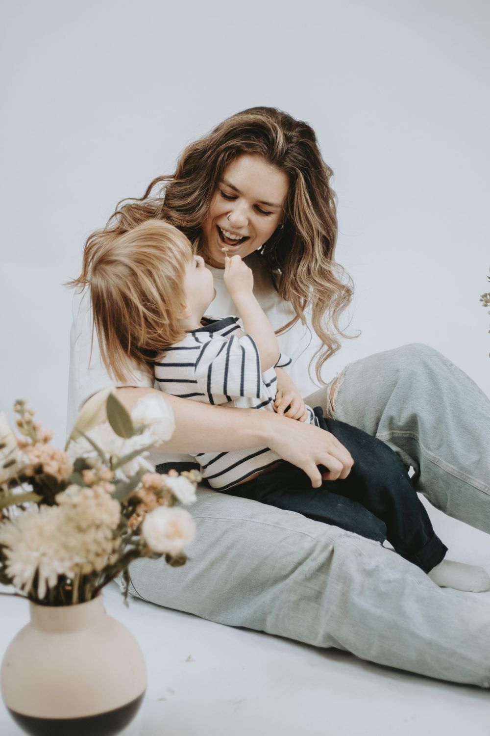 A joyful mother and young child laughing and playing together on the floor beside a floral vase in a bright studio.