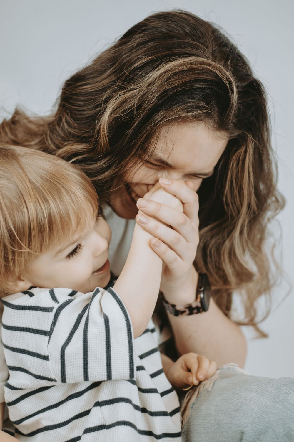 A mother with wavy brown hair kisses her toddler's hands while the child looks up at her lovingly.