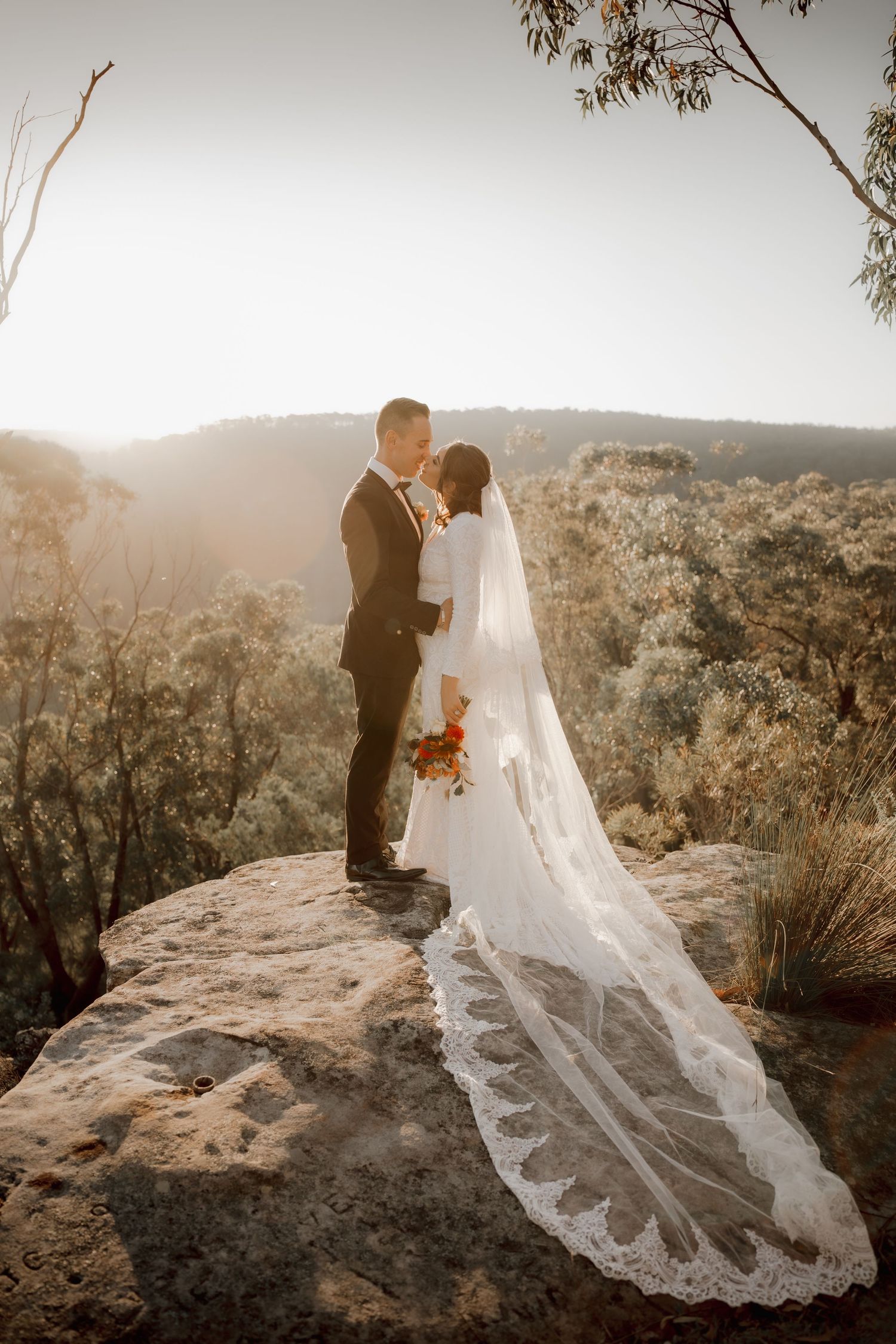 Bride and groom stand on rocky cliff overlooking lush Australian bushland at golden hour, her lace veil trailing beautifully.