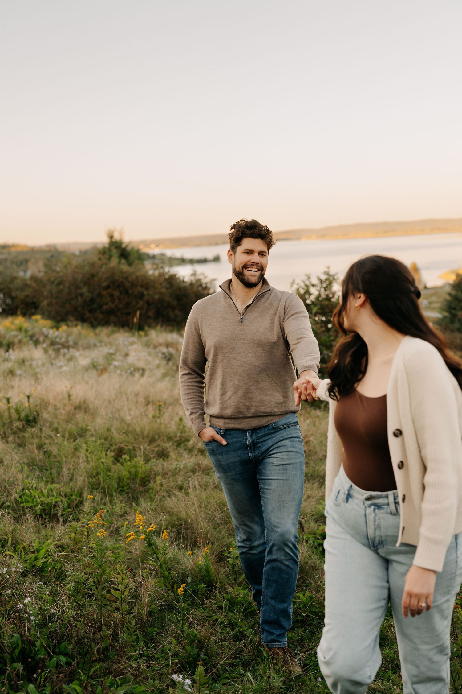 Smiling couple holding hands while walking through a grassy field at golden hour near a scenic waterfront in Laurencetown.