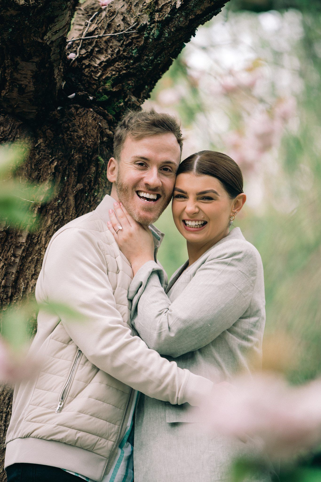 Happy couple embracing and smiling beside a tree with pink blossoms in a lush green garden.