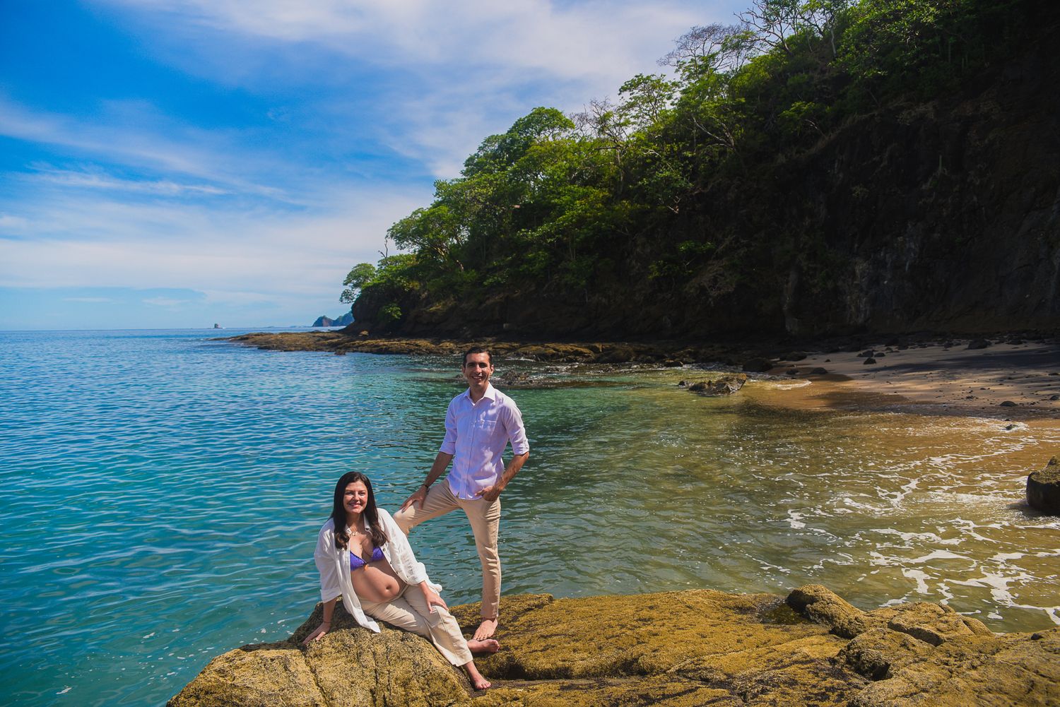 Couple posing on rocky shore with turquoise ocean and lush green cliffs in a tropical beach setting.