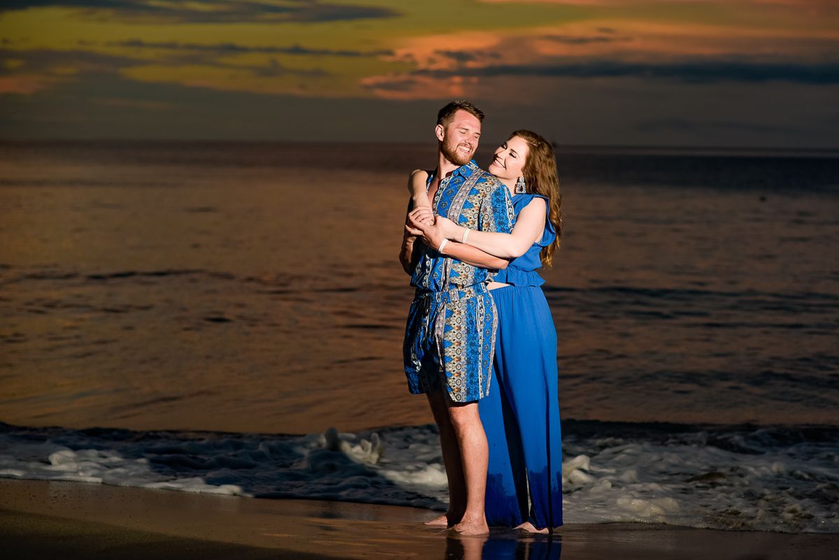 Couple embracing on a beach at sunset, wearing blue outfits, with ocean waves and colorful sky behind them in Tamarindo area