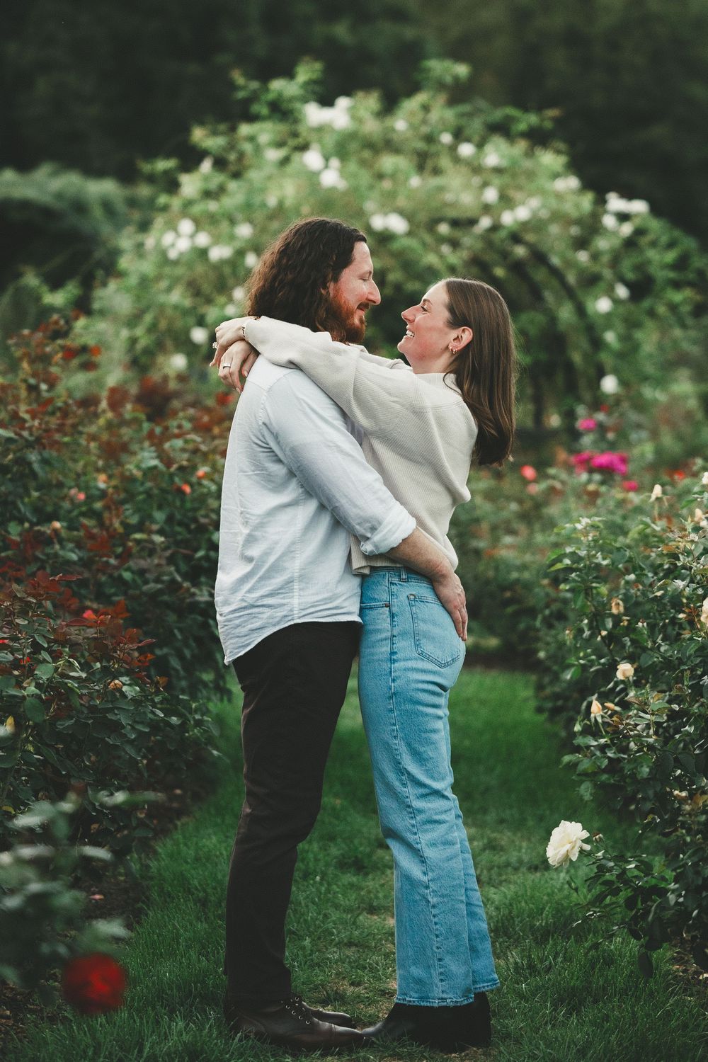 Couple embracing and laughing in a lush rose garden with colorful blooms and a floral arch backdrop.