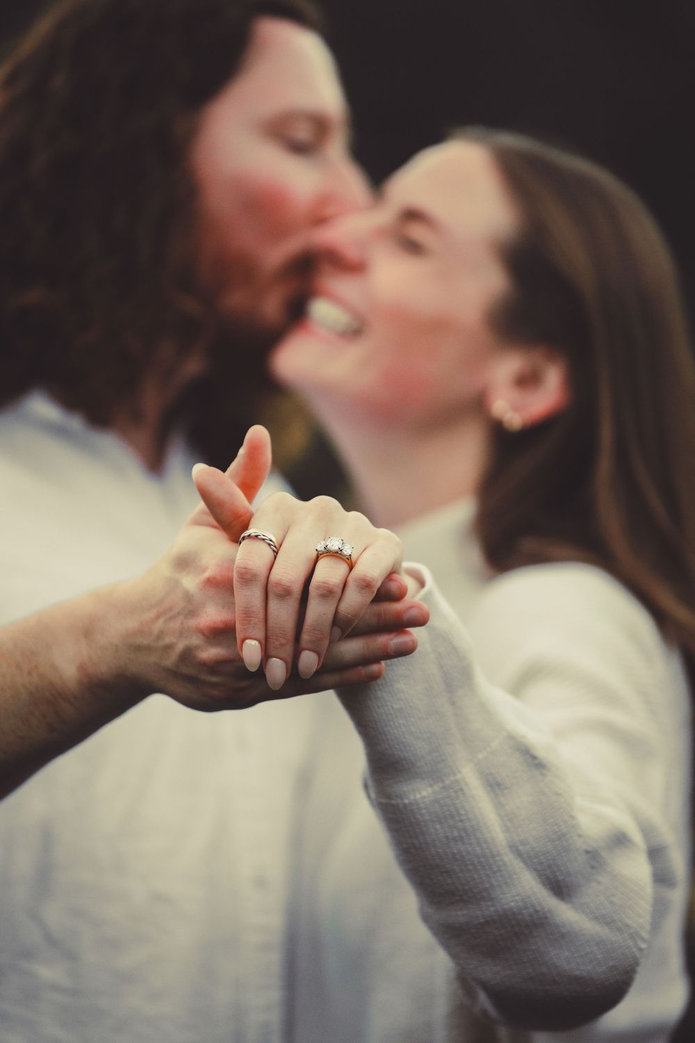 Couple sharing a kiss while holding hands showing engagement rings, woman smiling in white sweater, romantic proposal moment.