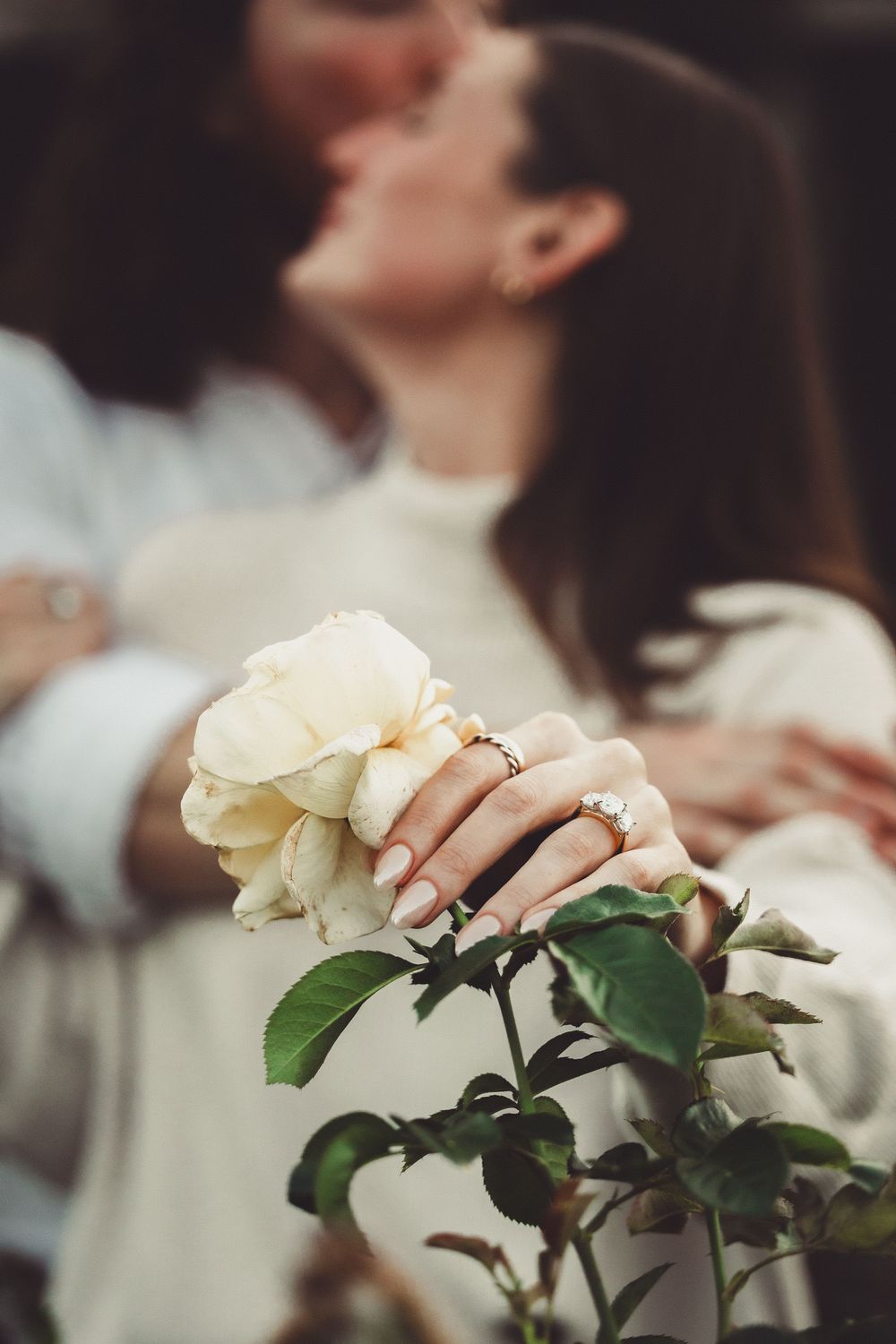 Couple kissing behind a white rose bush, with an engagement ring visible on a woman's hand.