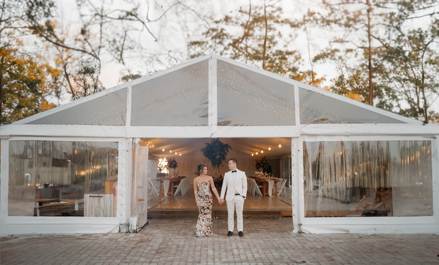 Couple in formal attire standing before a large elegant transparent wedding tent with string lights at dusk.