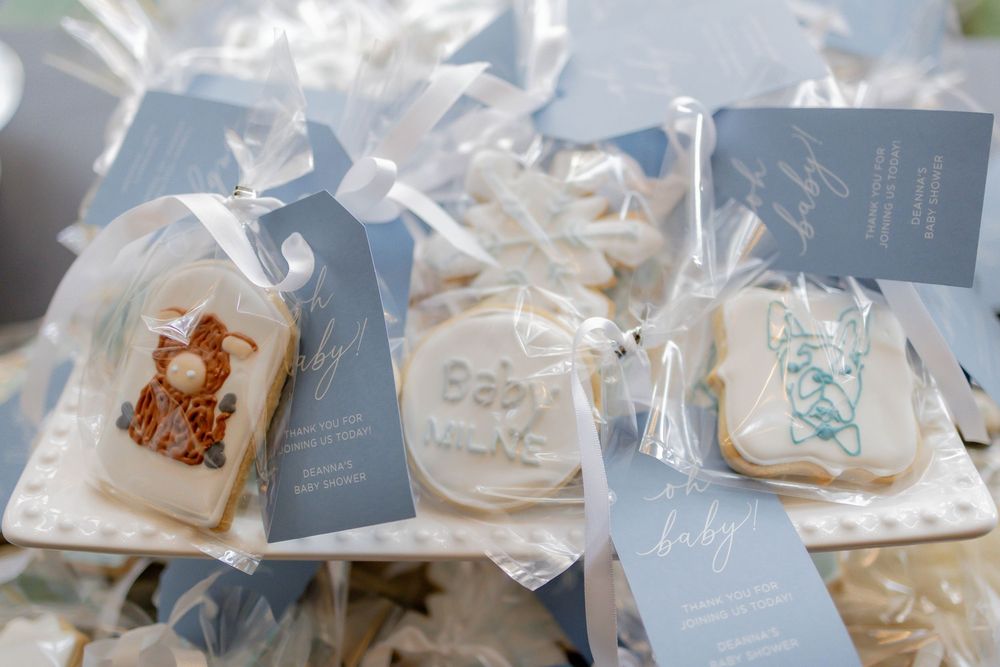 Baby shower sugar cookies with blue tags, wrapped in cellophane, featuring teddy bear and baby designs on a white tray.