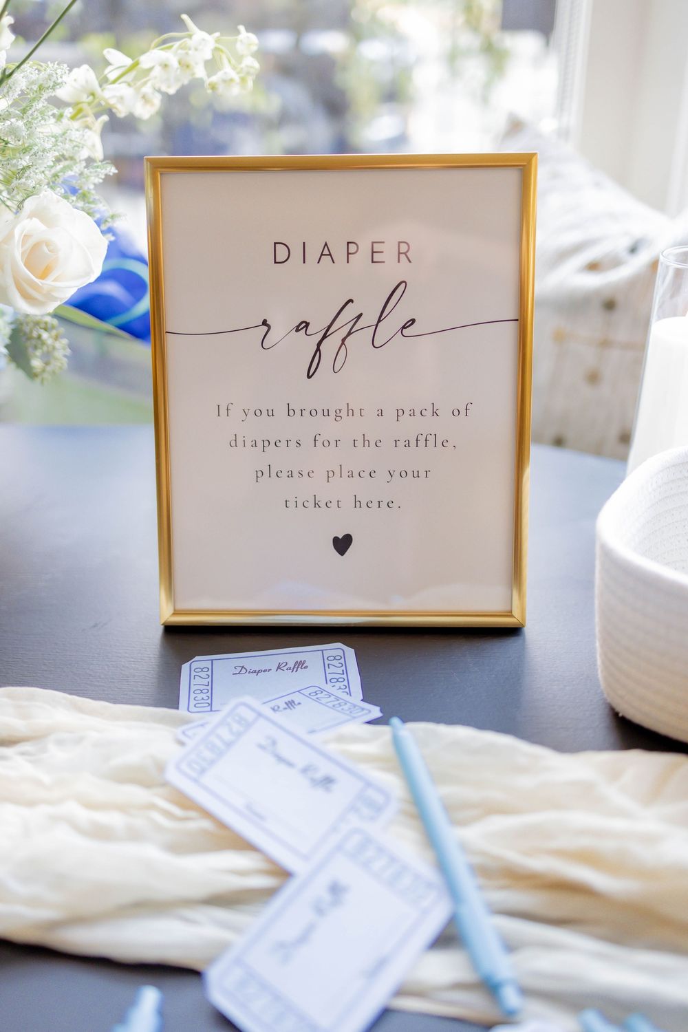 Gold framed diaper raffle sign at baby shower with blue raffle tickets and pen on dark table.